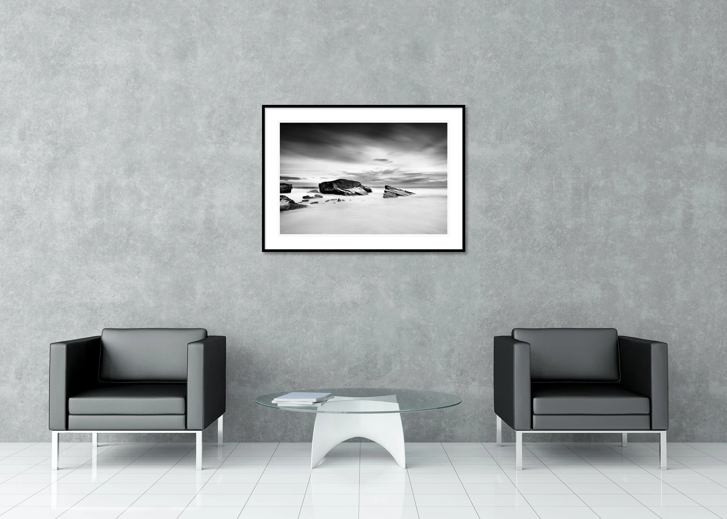 Monochrome long-exposure seascape print in a classic frame with passe-partout showing a rocky coast with large rocks emerging from smooth water under overcast clouds