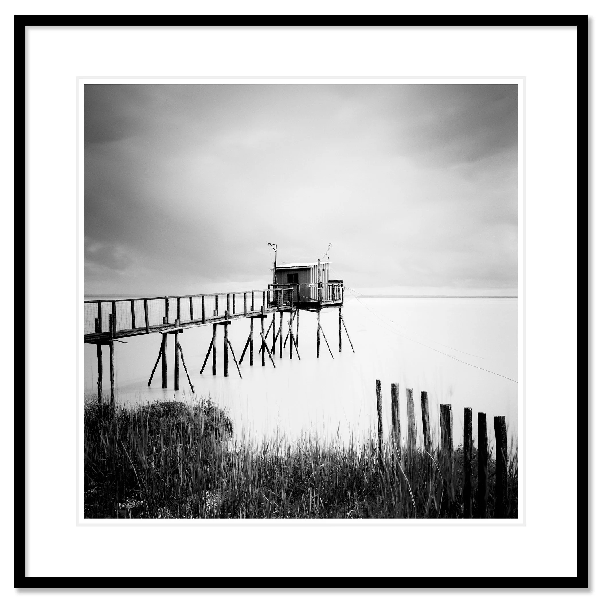 Gerald Berghammer - Black and white seascape photography. Small wooden hut on stilts connected by a long walkway over water with grass and a cloudy sky. Classic framed black