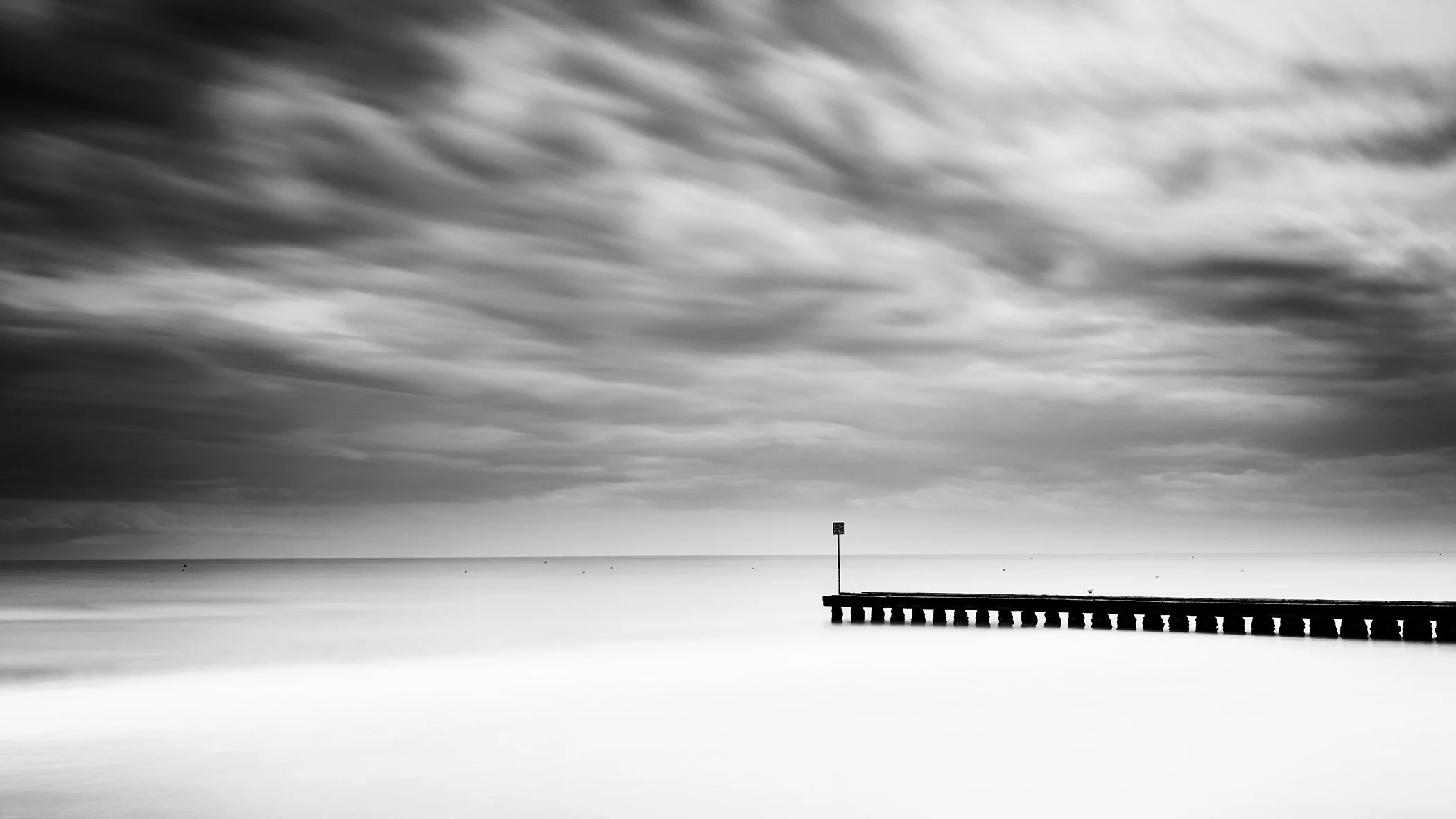 Black and white long-exposure photograph of a calm sea with dramatic clouds and a wooden groyne extending from the right.