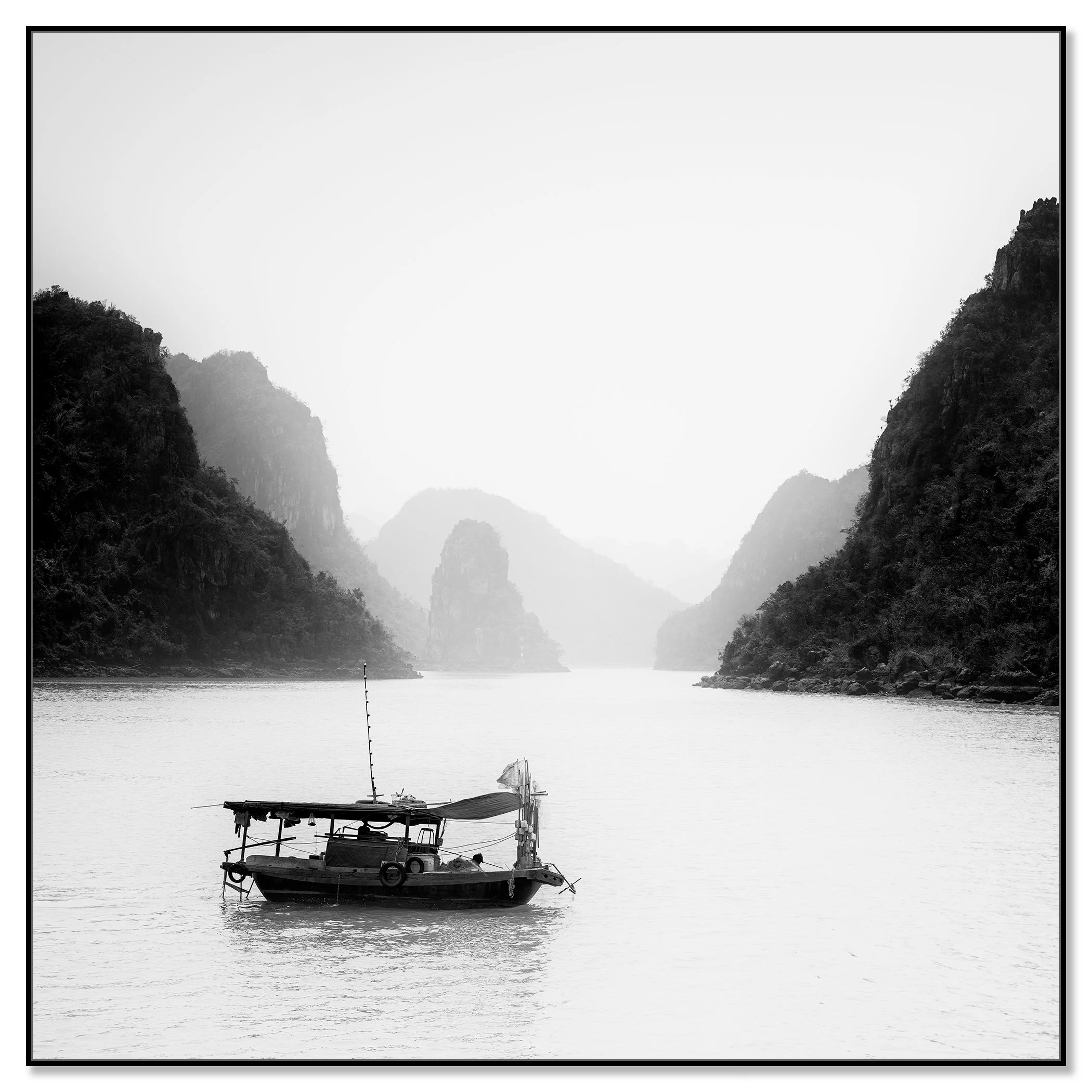 Black-and-white photograph of a fishing boat on calm water between misty limestone mountains – framed ArtBox black