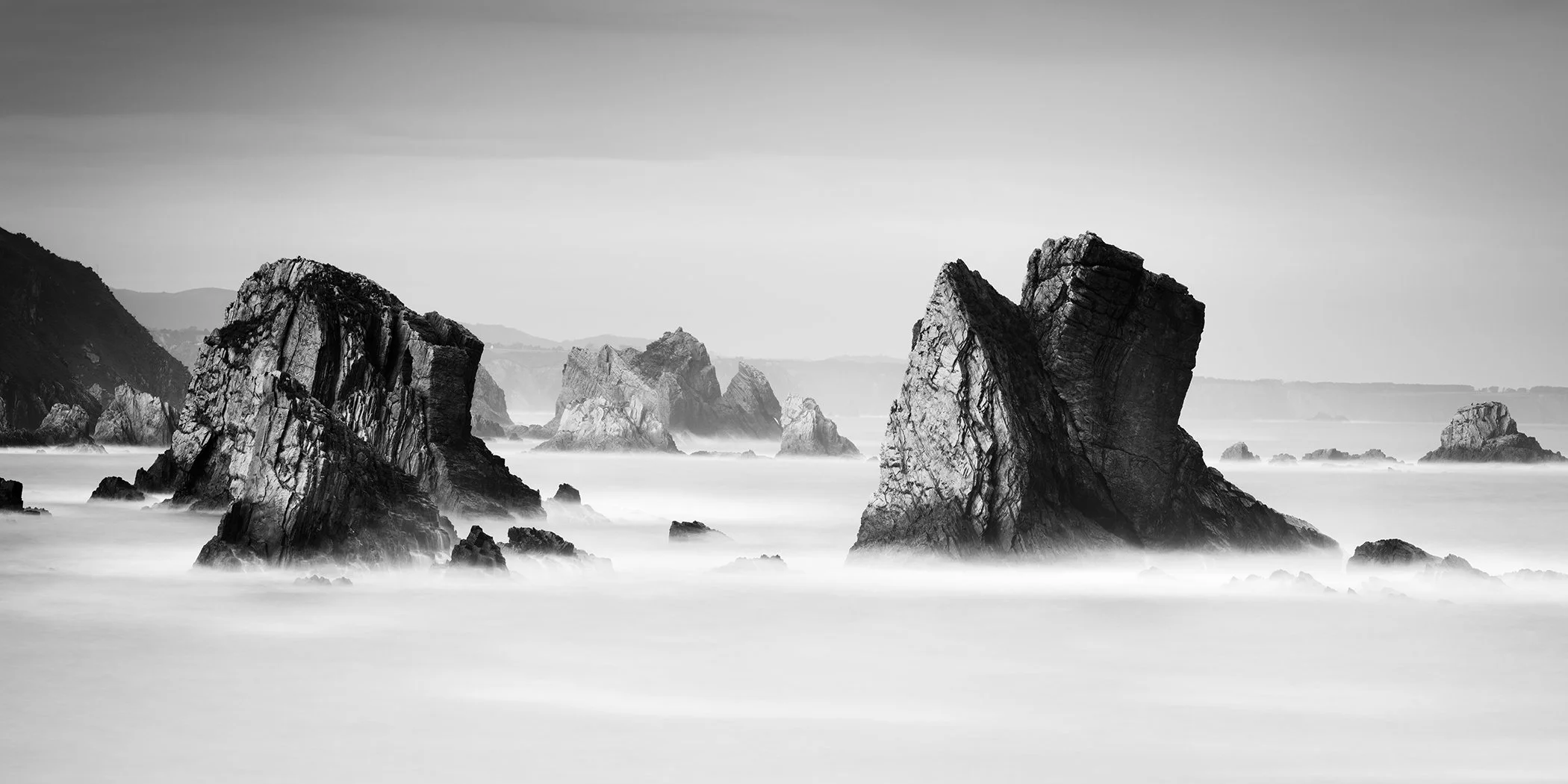 Black-and-white photograph of rocky sea stacks with misty waves along a rugged coastline.