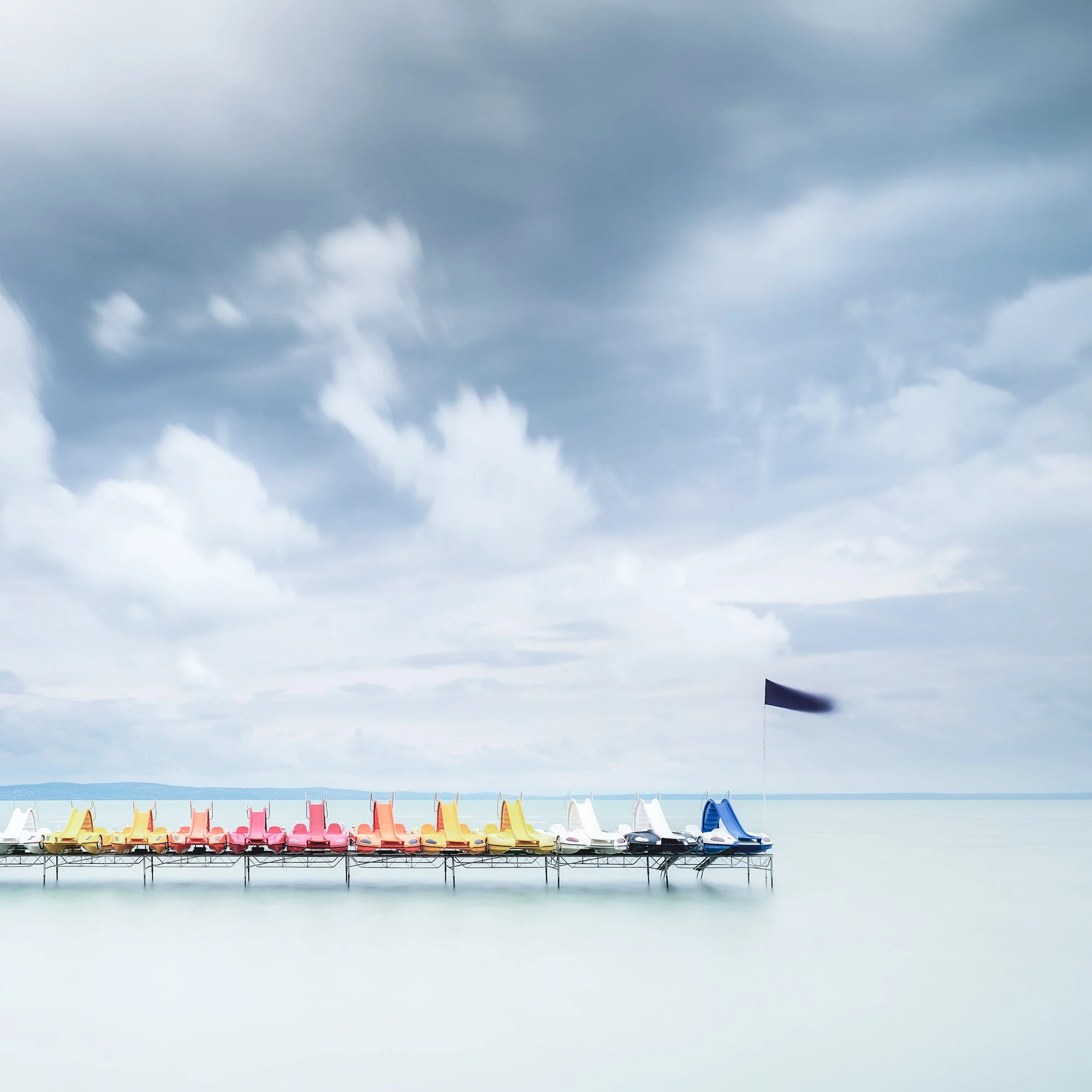 Gerald Berghammer - Color long exposure waterscape photography. Colorful paddle boats lined up on a dock over calm water, under a cloudy sky. Print detail 2