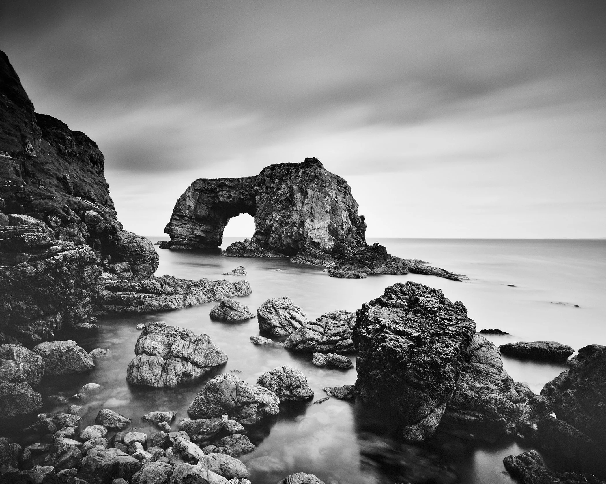 Great Pollet Sea Arch in black and white, with rugged coastal rocks, smooth water, and cloudy sky.
