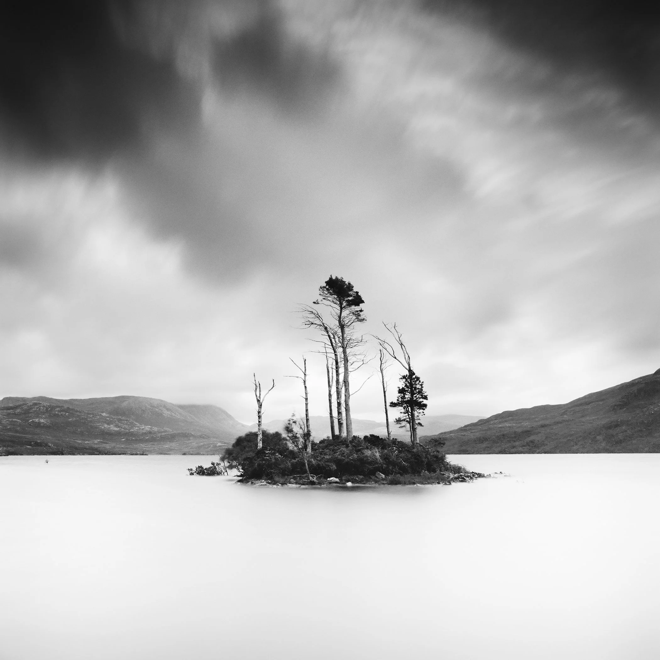 © 2012 Gerald Berghammer - Black and white waterscape photography. Small island with tall, thin trees surrounded by water and distant mountains under a cloudy sky.