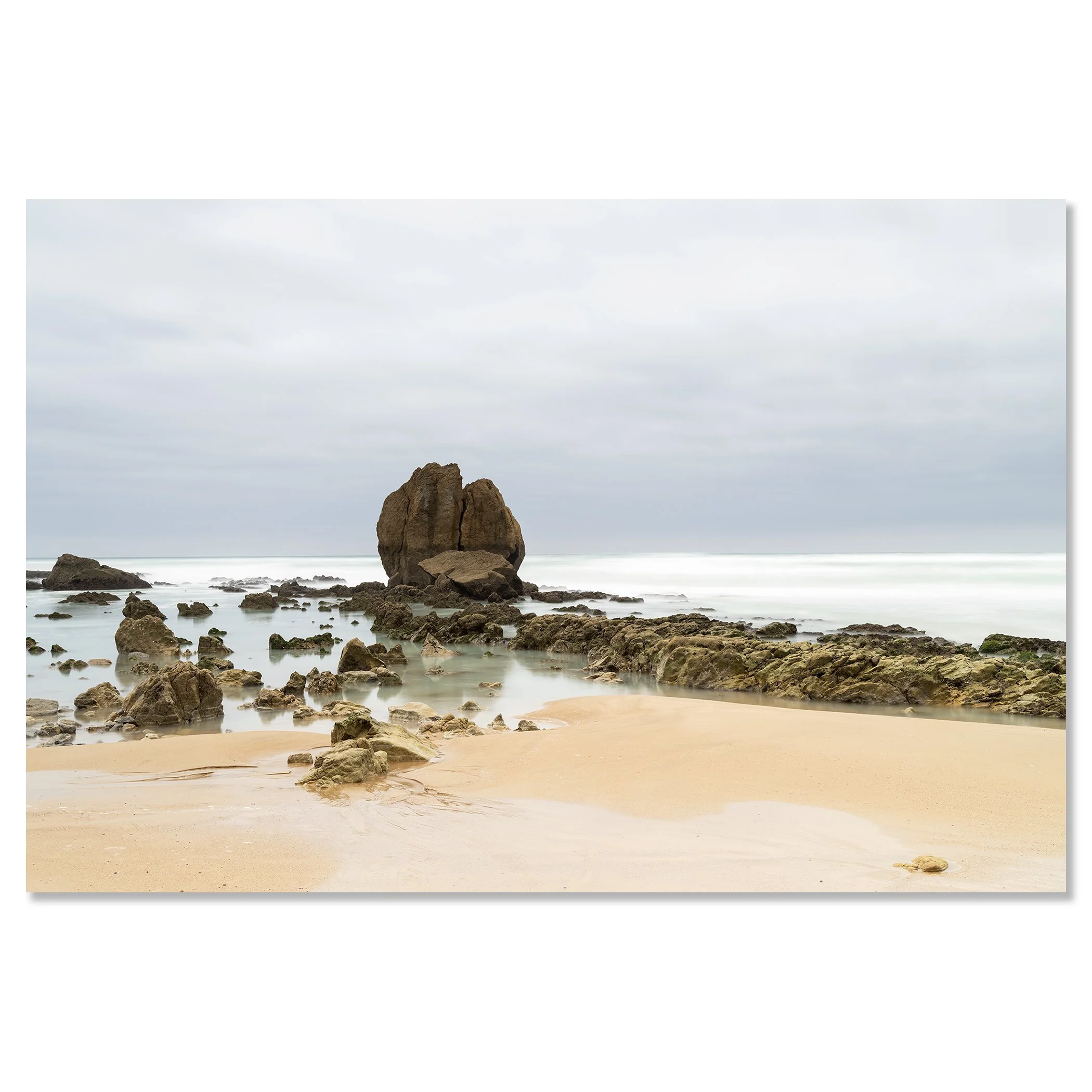 Gerald Berghammer - Color Seascape Photography. A beach with rocks and sand, with large rock formations rising from the water, under a cloudy sky. Chromaluxe frameless