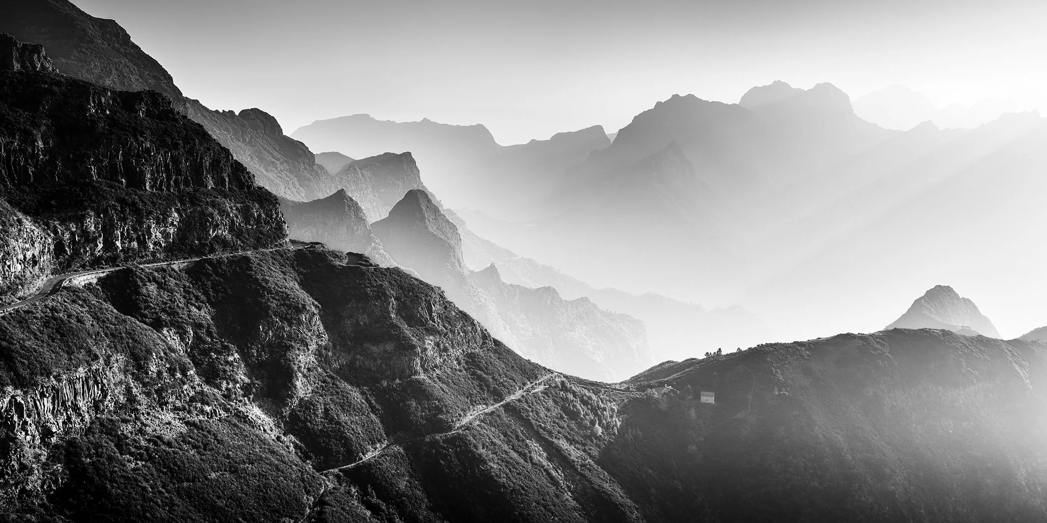 Black-and-white mountain landscape featuring layered peaks, a winding road and soft atmospheric haze.