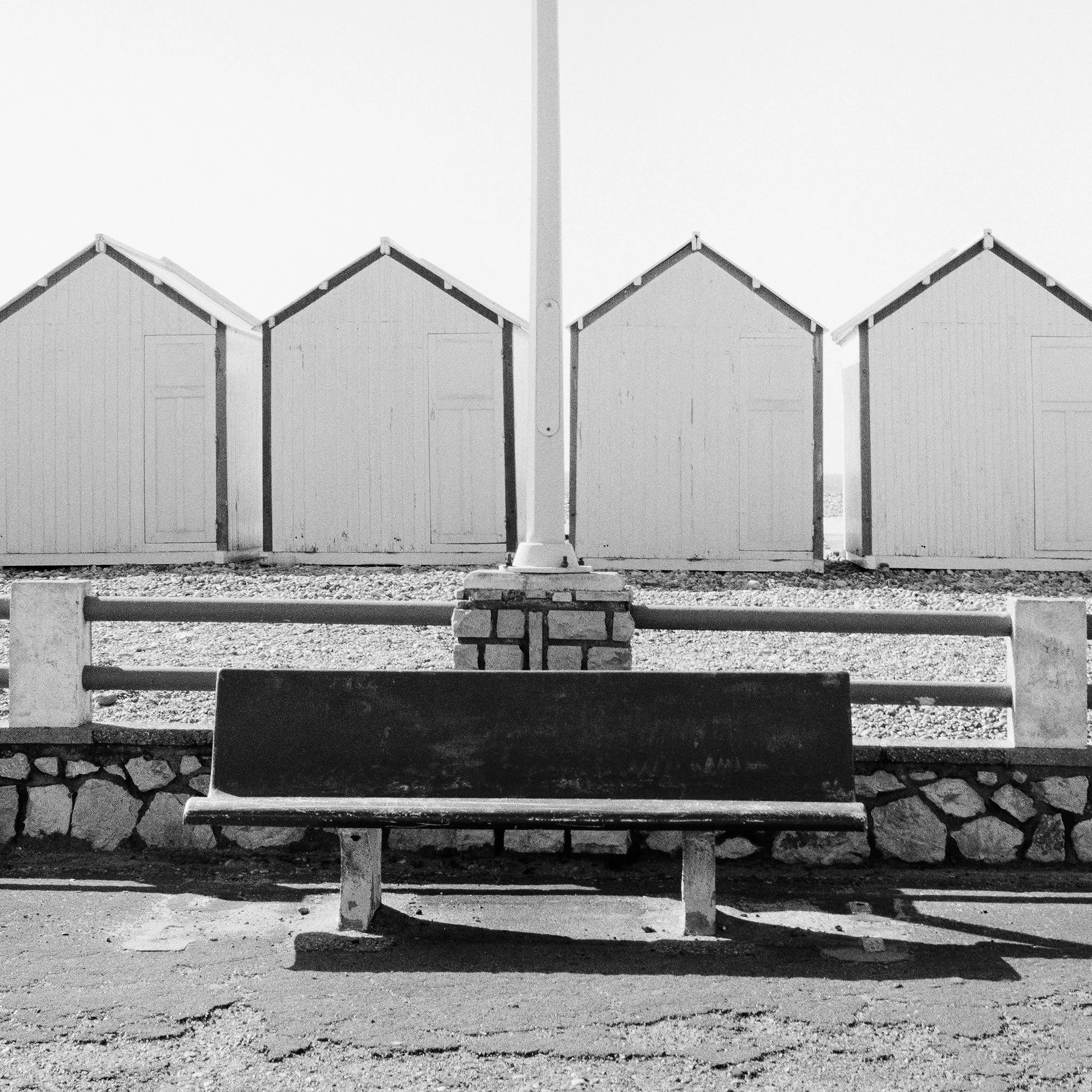 Close view of Bench on the Promenade Study 1 with a centred streetlamp, empty bench and repeating beach huts by Gerald Berghammer.