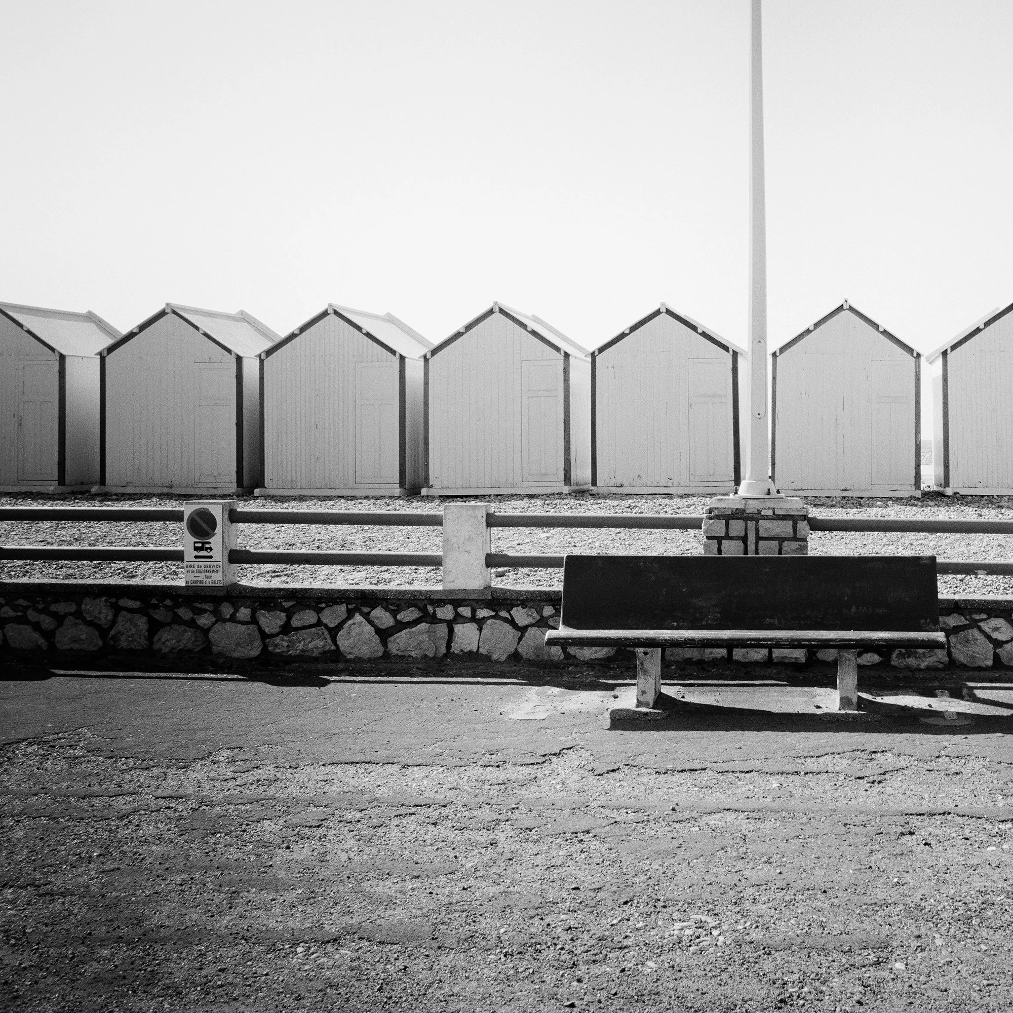 Empty bench on the promenade facing beach huts, centred streetlamp, minimalist black-and-white coastal photograph, Detail 1