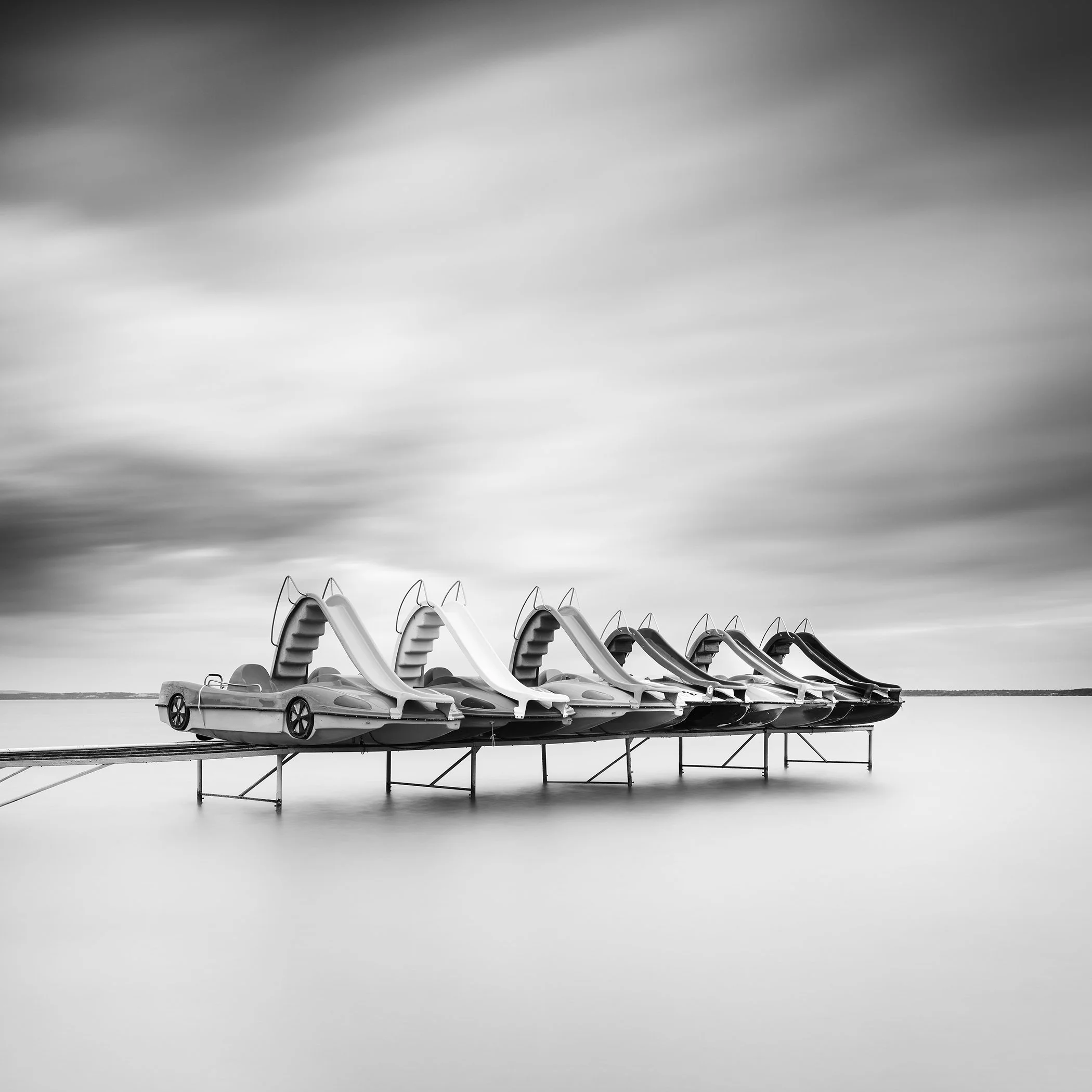 Gerald Berghammer - Black and white long exposure waterscape photography. Five pedal boats on a platform over water, with a cloudy sky in the background.