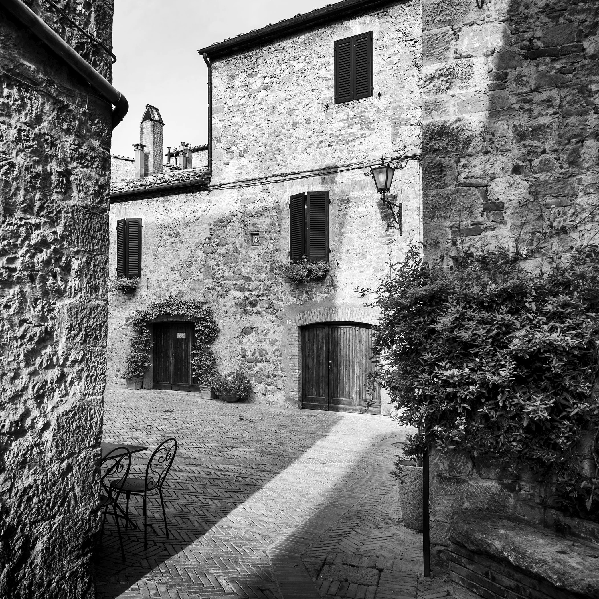 Gerald Berghammer - Black and white cityscape photography. Old stone building with wooden shutters, potted plants and outdoor seating in a narrow courtyard.