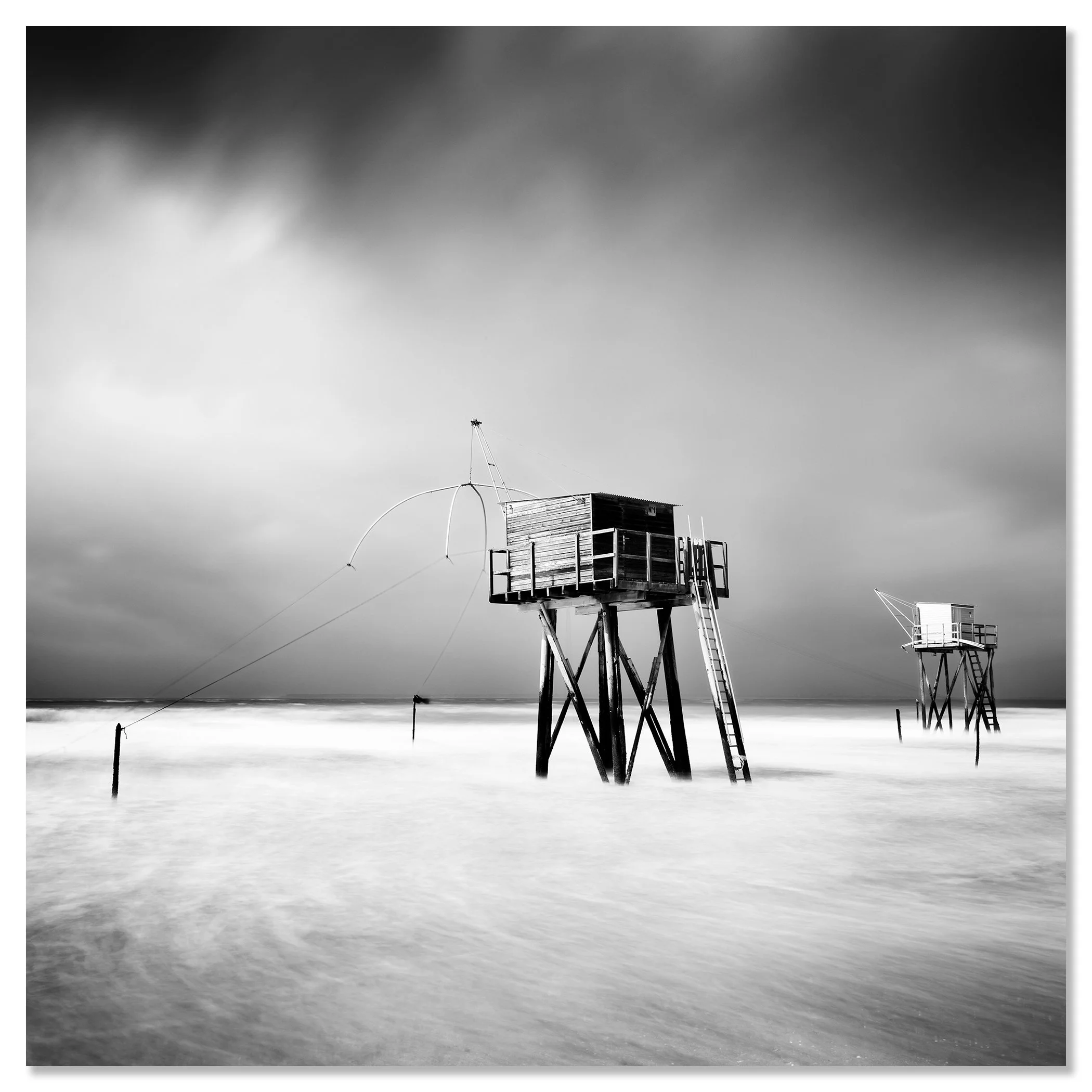 Black and white long-exposure photo of wooden fishing huts on stilts, reached by ladders, over calm sea water under a dramatic cloudy sky – dibond frameless