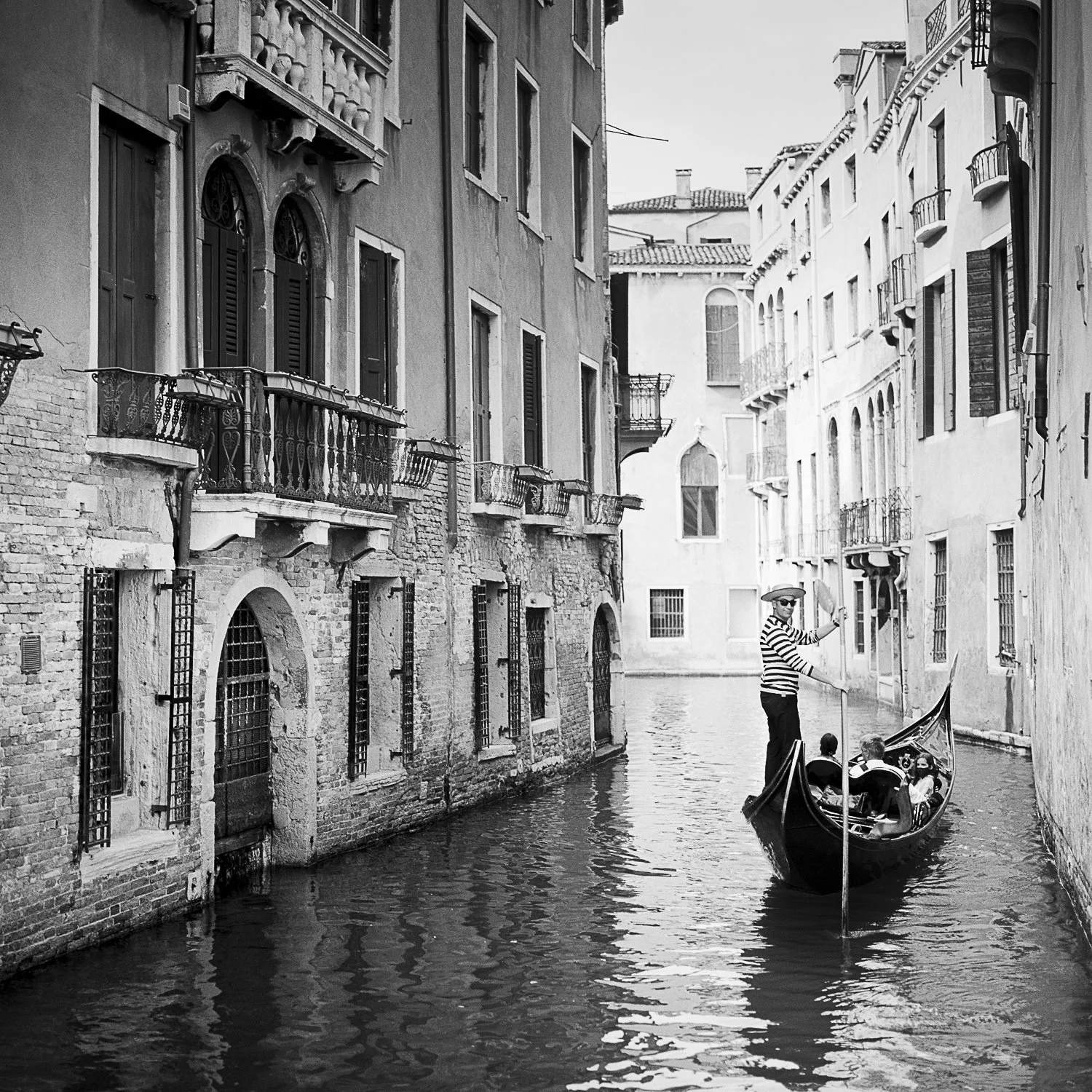 Black-and-white photograph by Gerald Berghammer showing a gondolier steering through a narrow Venetian canal lined with historic buildings.