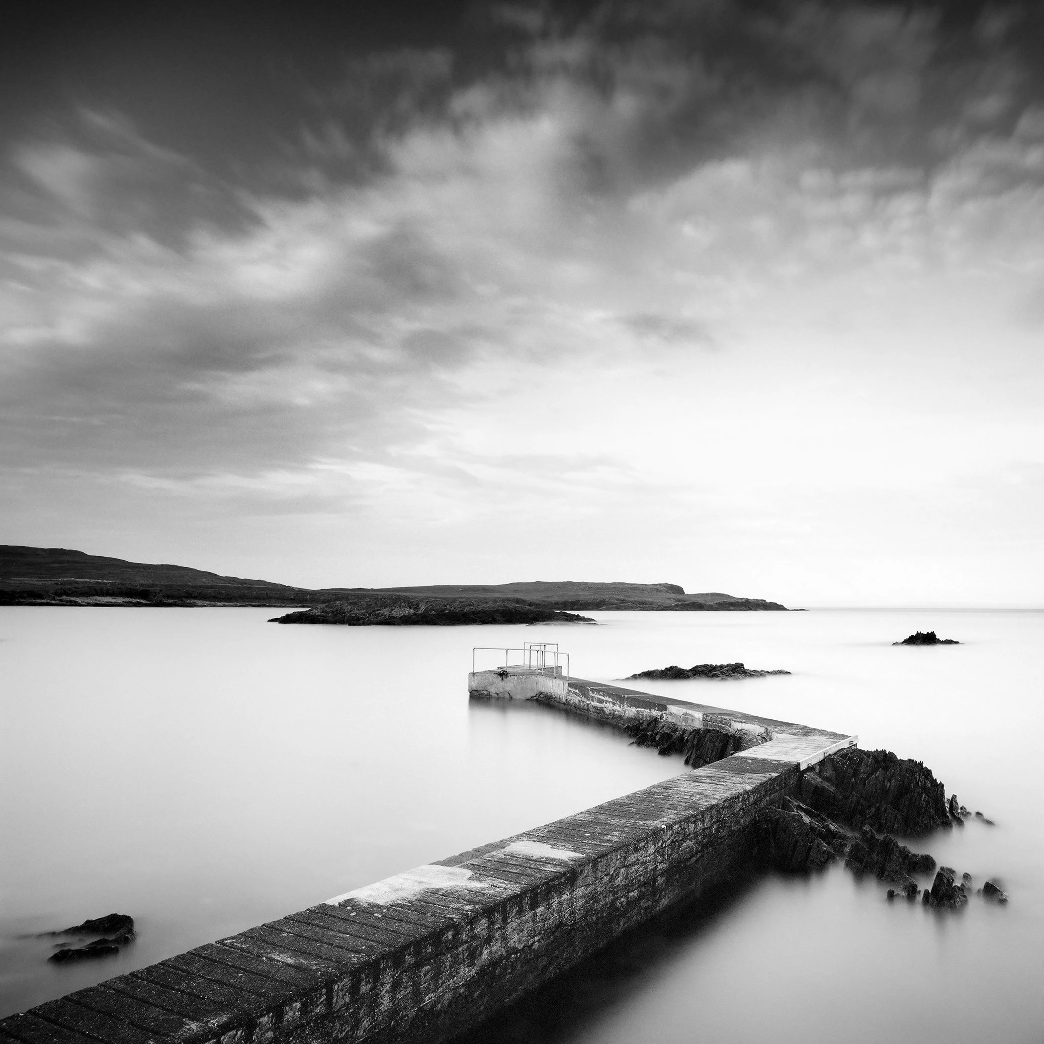 © 2014 Gerald Berghammer - Black and white seascape photography. A Concrete pier extends into calm seawater with rocky edges, under a cloudy sky.