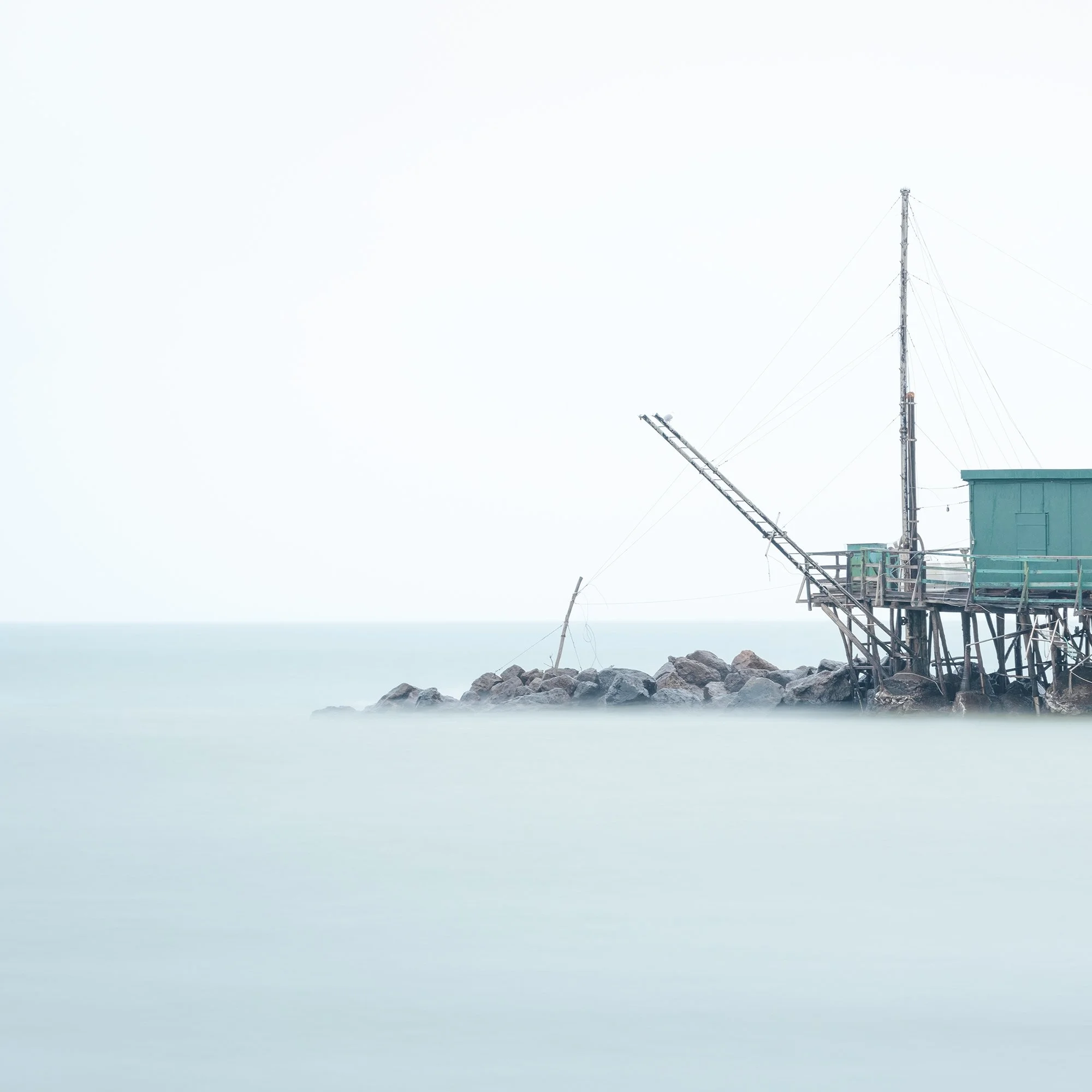 © 2024 Gerald Berghammer - Color minimalist photography. A small house built on stilts over water on rocks, with a fishing pole, in a minimalist, misty seascape. Print detail 1