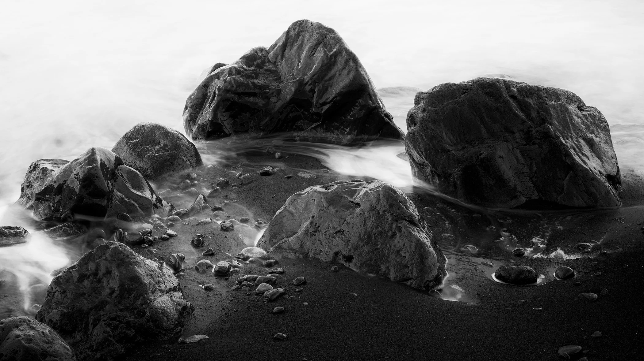 Black and white seascape with large rocks on wet sand and soft waves washing around them