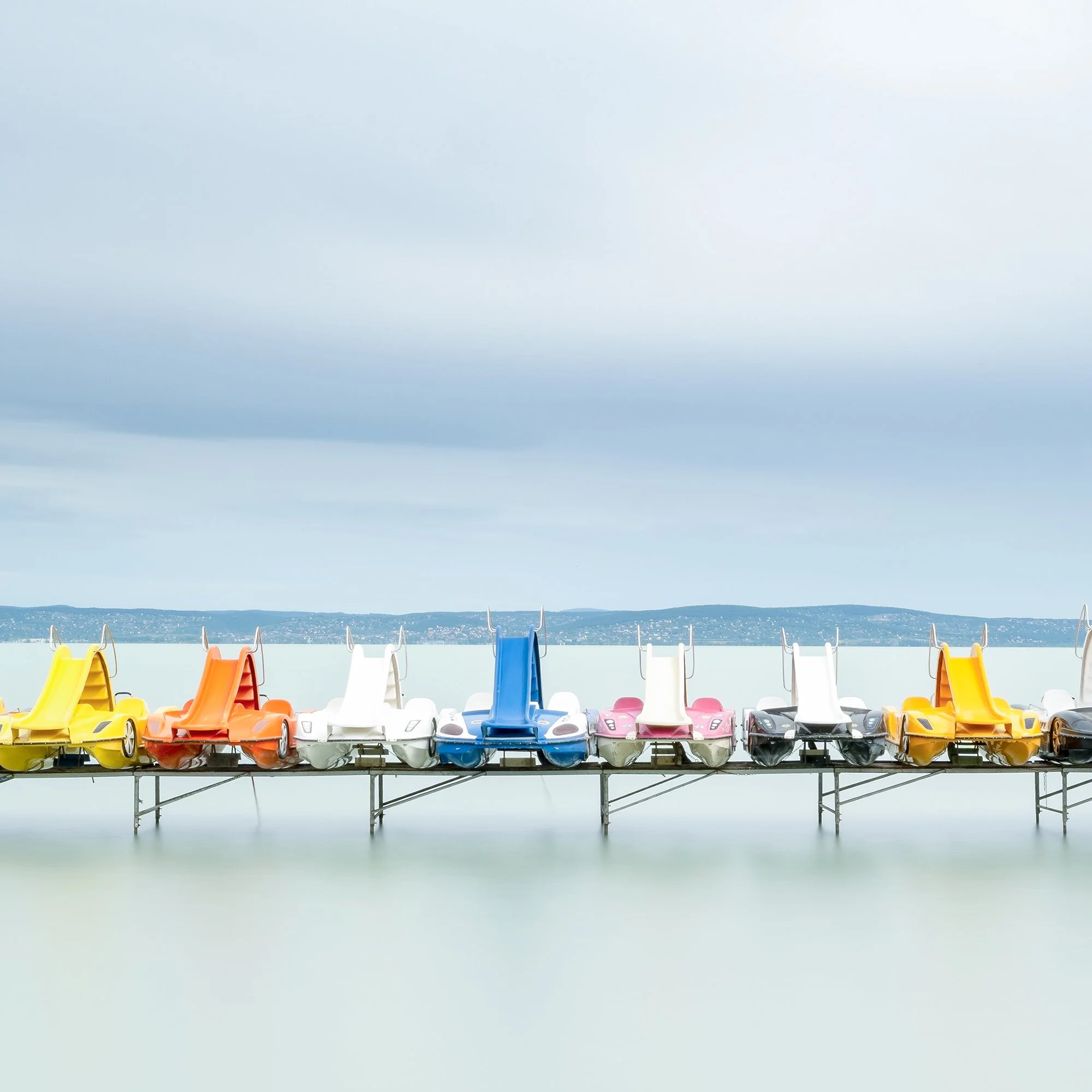 Gerald Berghammer - Color waterscape photography. Colorful pedal boats and a catamaran lined up on a dock on a calm body of water under a cloudy sky. Print detail 3