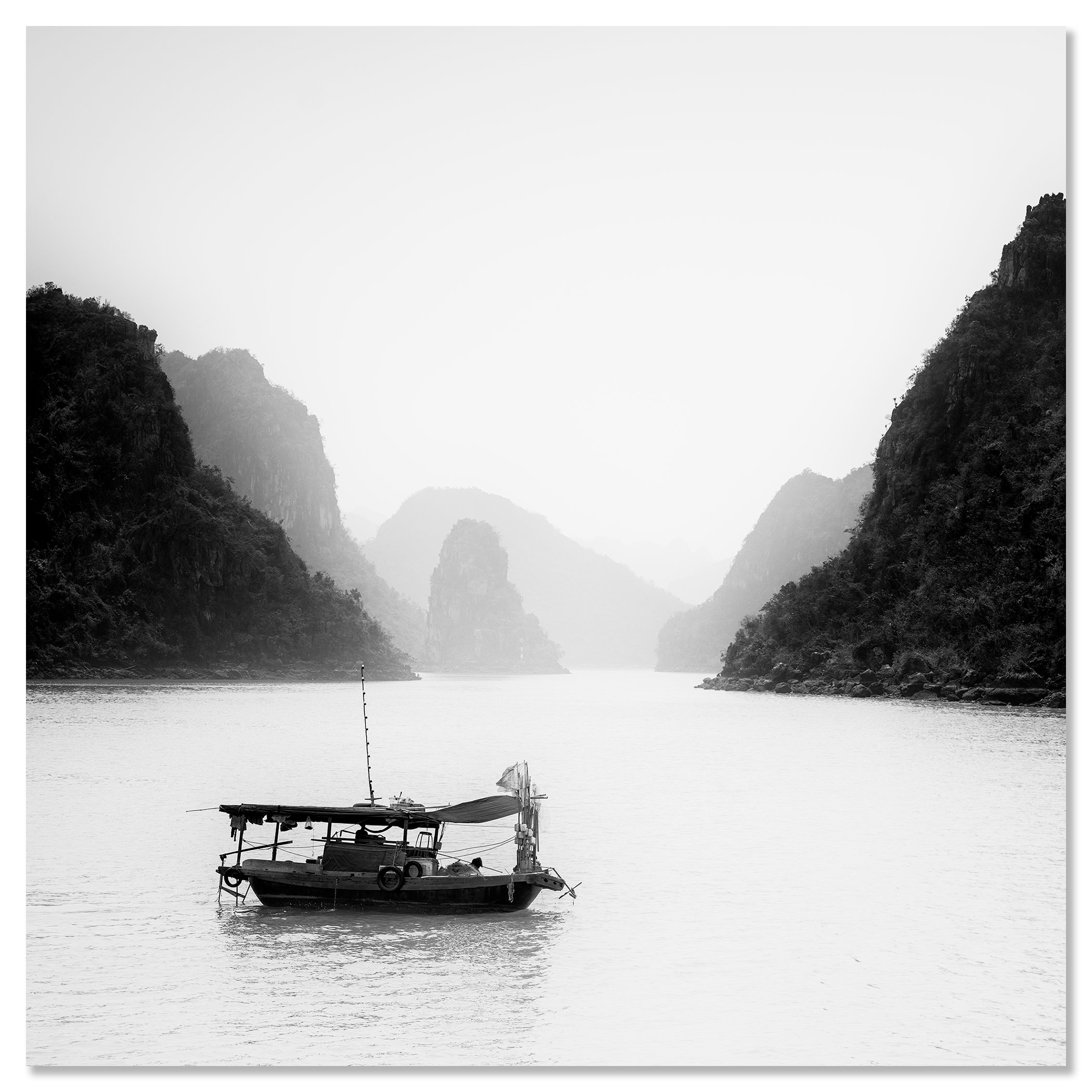 Black-and-white photograph of a fishing boat on calm water between misty limestone mountains – dibond frameless