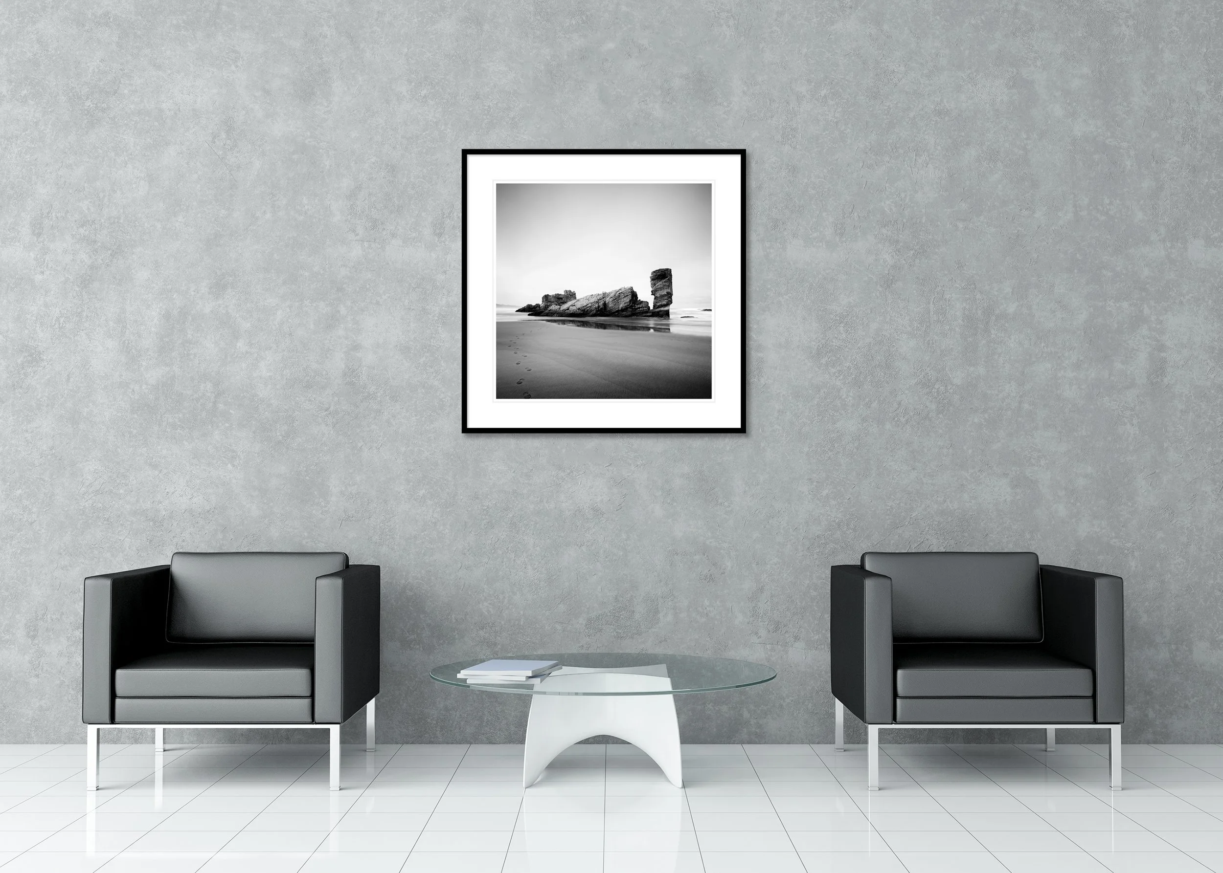 Black-and-white seaside view with damp sand, faint footprints, and a towering shore rock reflected in calm shallow water.