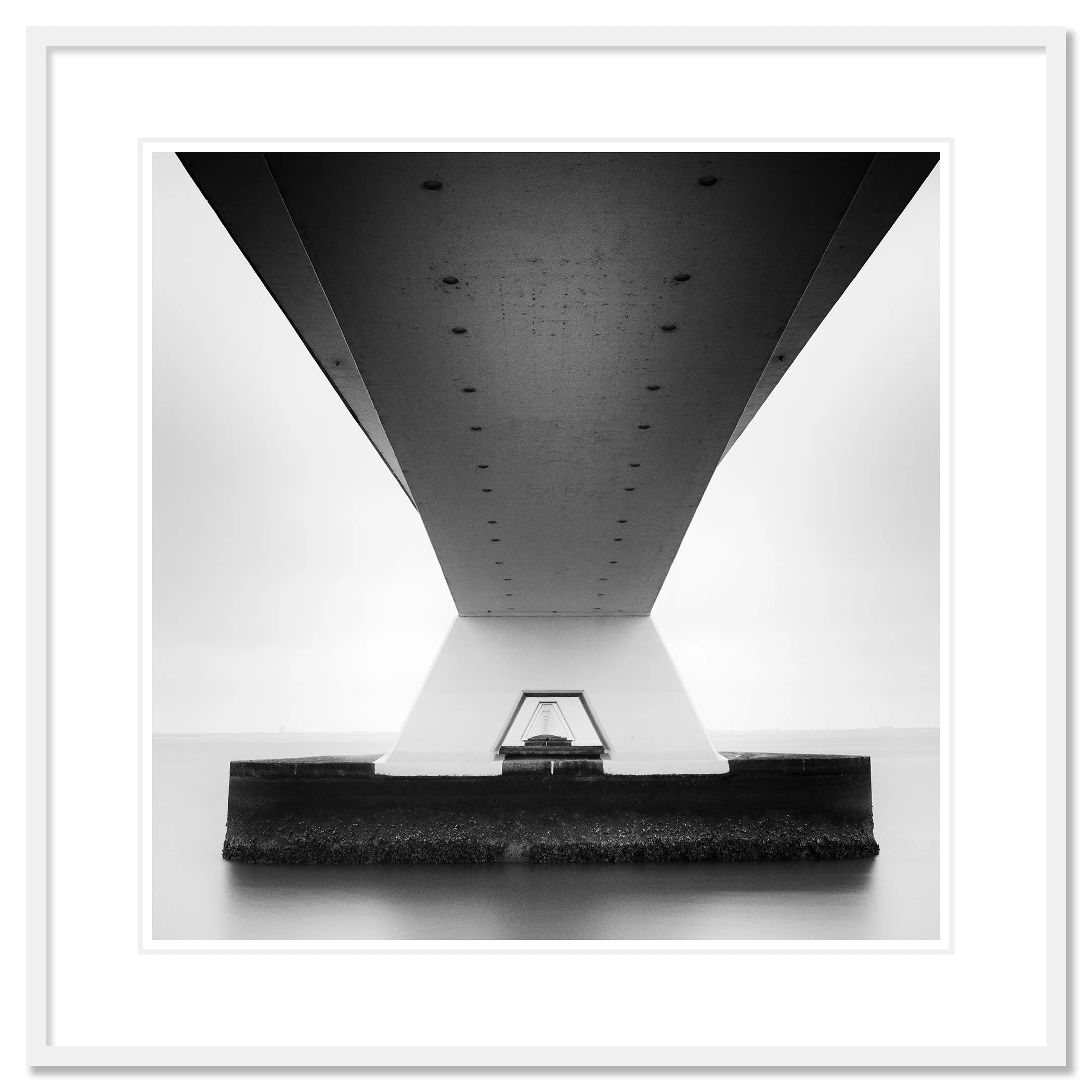 Gerald Berghammer - Black and white cityscape photo. Underneath view of a bridge with large support pillars, showing the underside and the water below. Classic framed white
