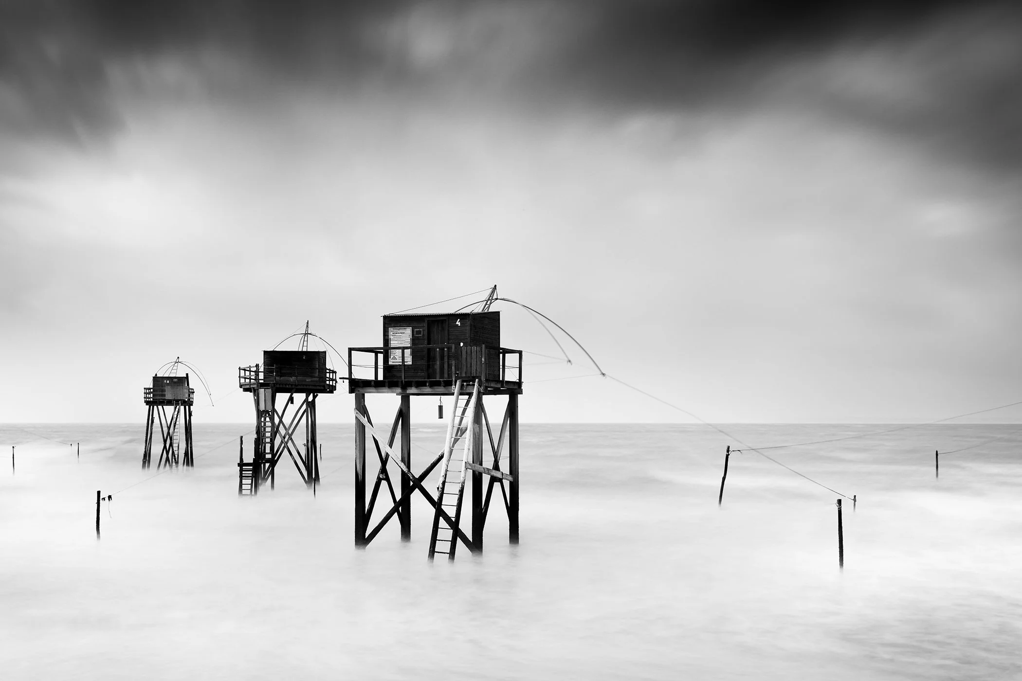 Black-and-white long-exposure seascape showing wooden fishing huts on stilts above soft, mist-like water.