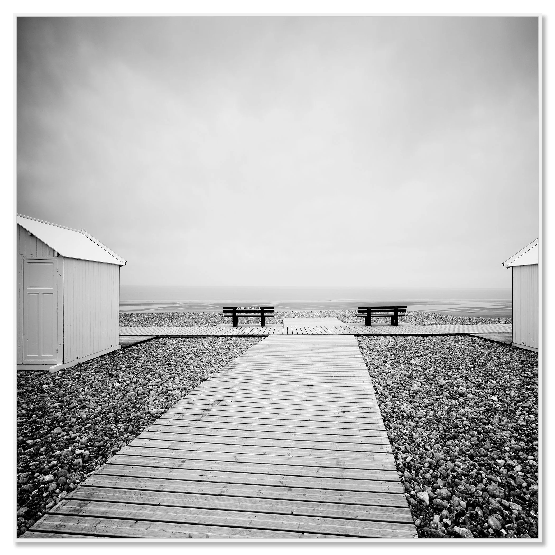 Black and white photo of a wooden boardwalk leading across a pebble beach to benches facing the sea – framed ArtBox white