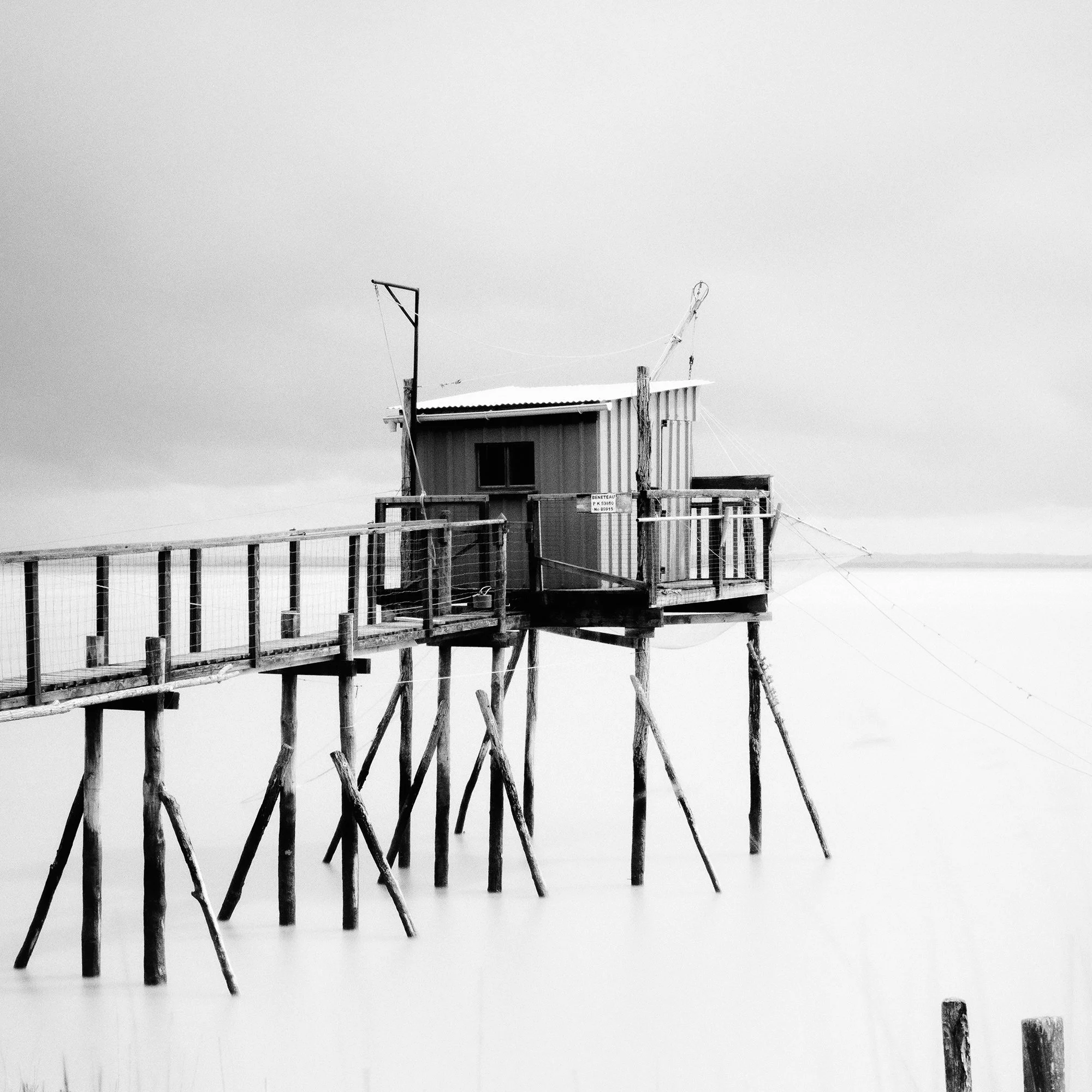 Gerald Berghammer - Black and white seascape photography. Small wooden hut on stilts connected by a long walkway over water with grass and a cloudy sky. Print detail 3