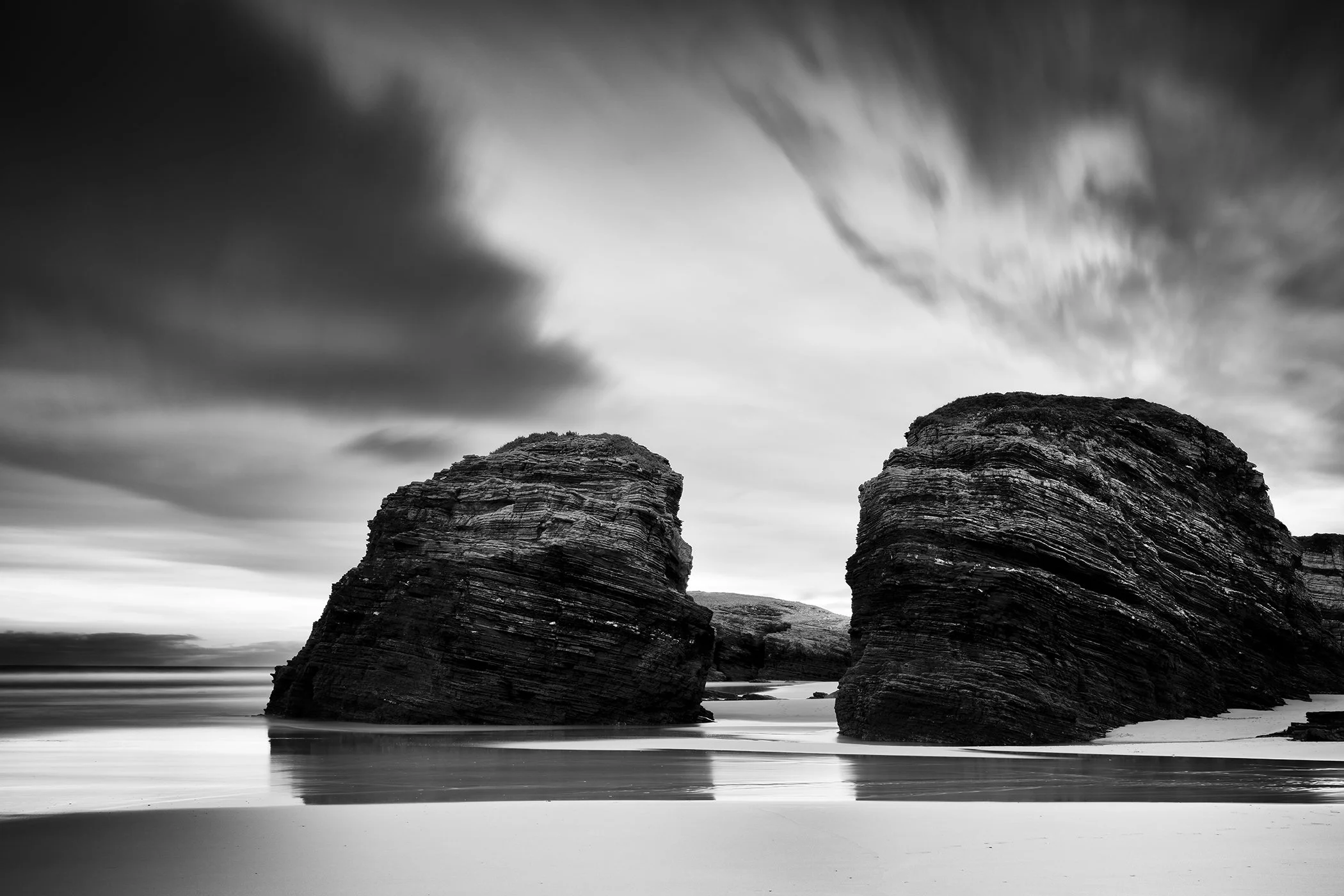 Black and white fine art landscape photograph of two rounded rock formations on a sea beach under a cloudy sky.