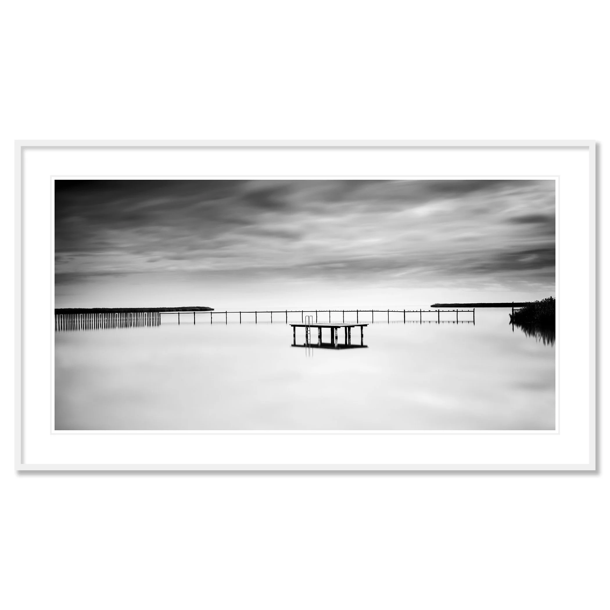 © 2023 Gerald Berghammer - Black and white minimalist waterscape. A calm body of water with a floating dock and a pier in the background, under a cloudy sky. Classic framed white