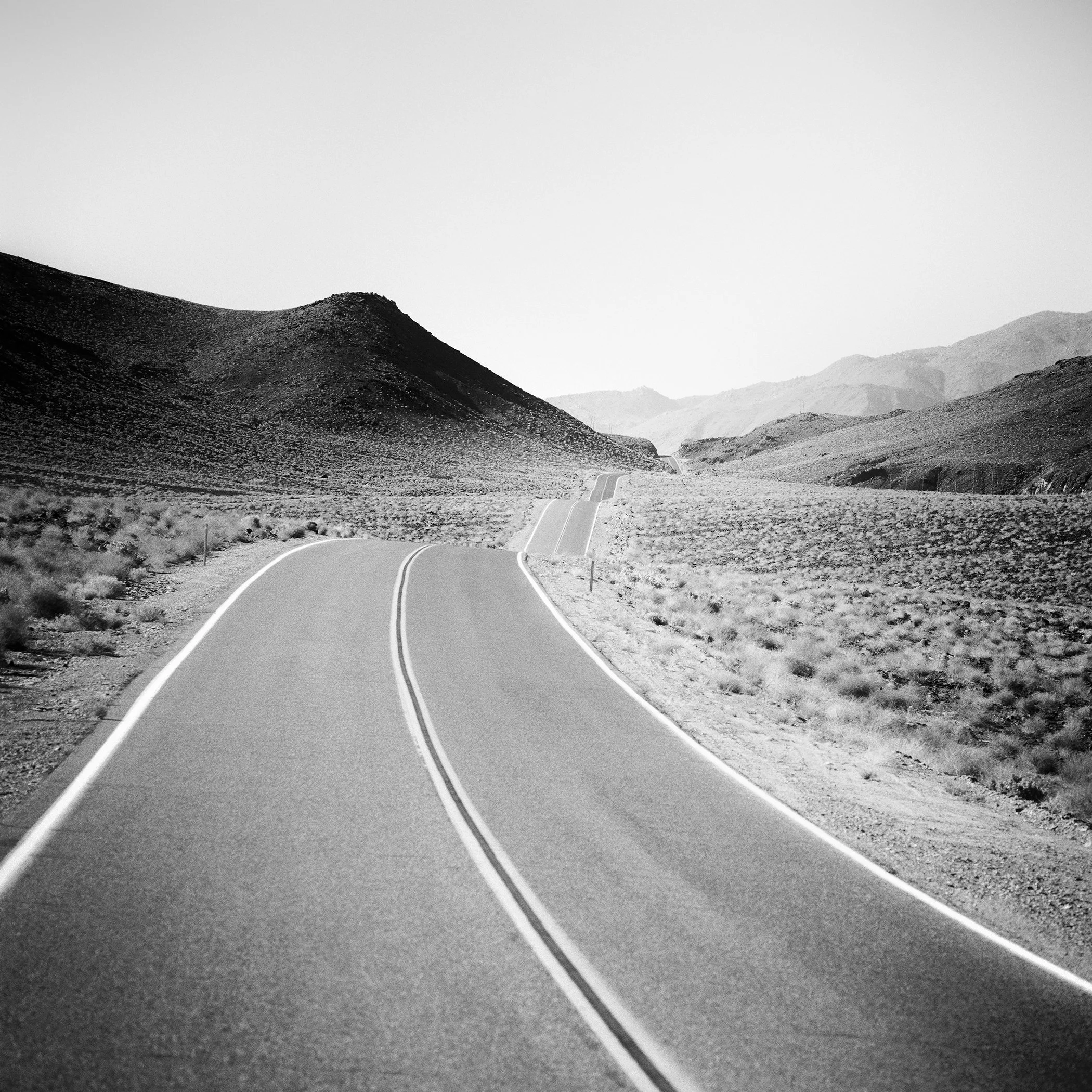 Minimalist black-and-white photograph of a winding desert road through rugged hills in Arizona, USA. Photo by Gerald Berghammer.