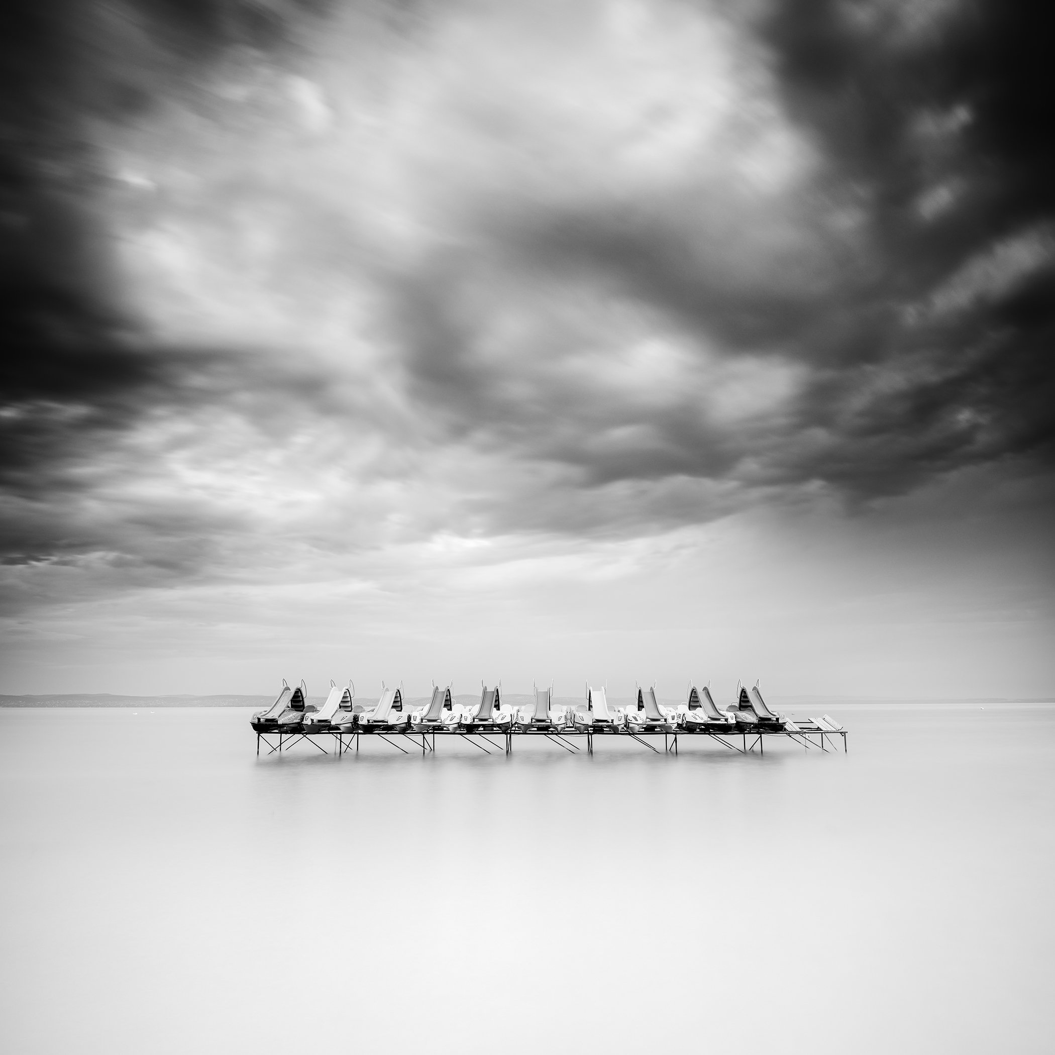 Fine art black-and-white photograph of pedal boats on a still lake beneath a brooding cloudy sky, by Gerald Berghammer