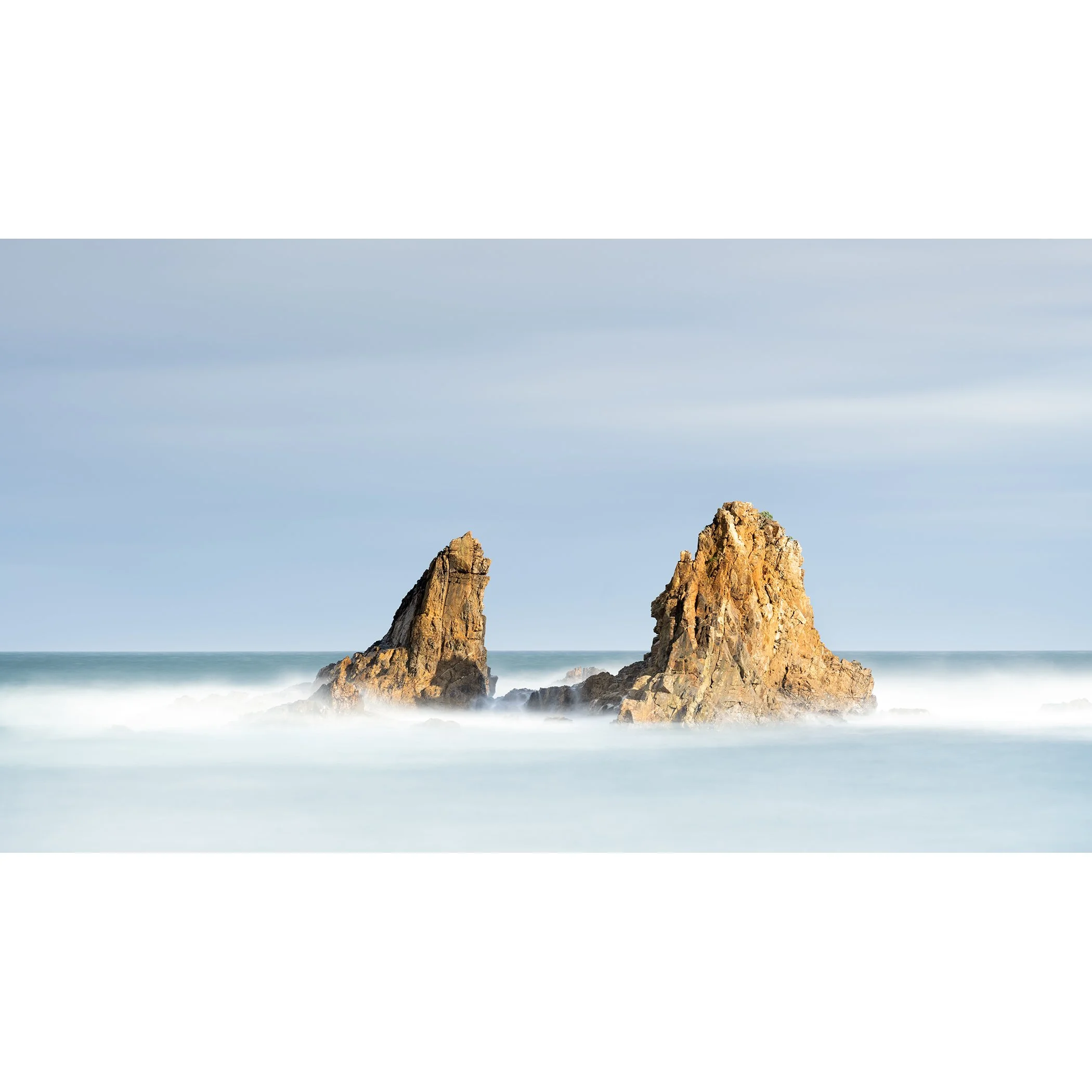 Gerald Berghammer - Color Seascape Photography. Two large rocks rising out of the ocean with waves crashing around them under a blue sky.
