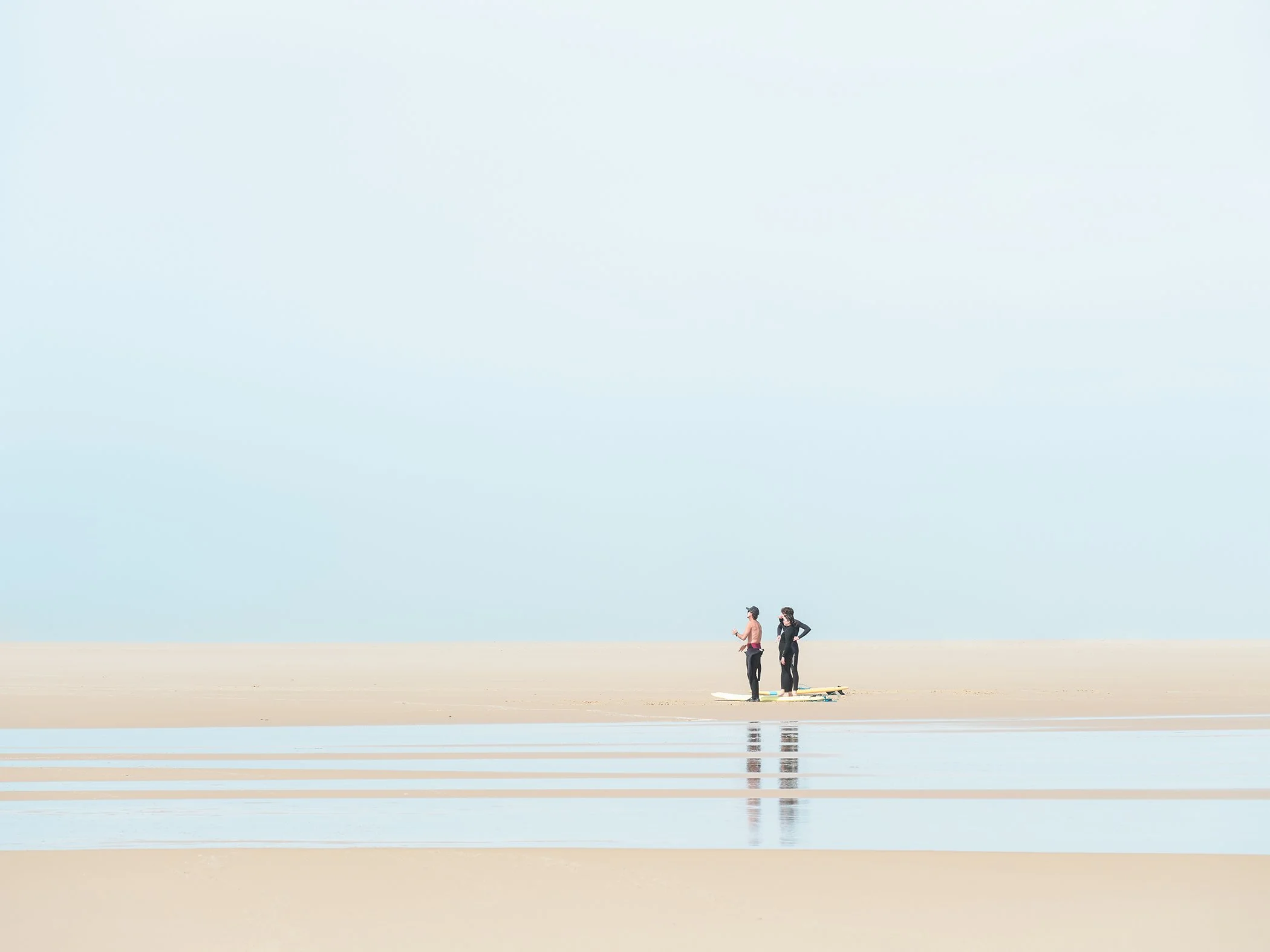 Three surfers on a vast minimalist beach in France with shallow tidal water, pale sky and calm reflections. Photo by Gerald Berghammer.