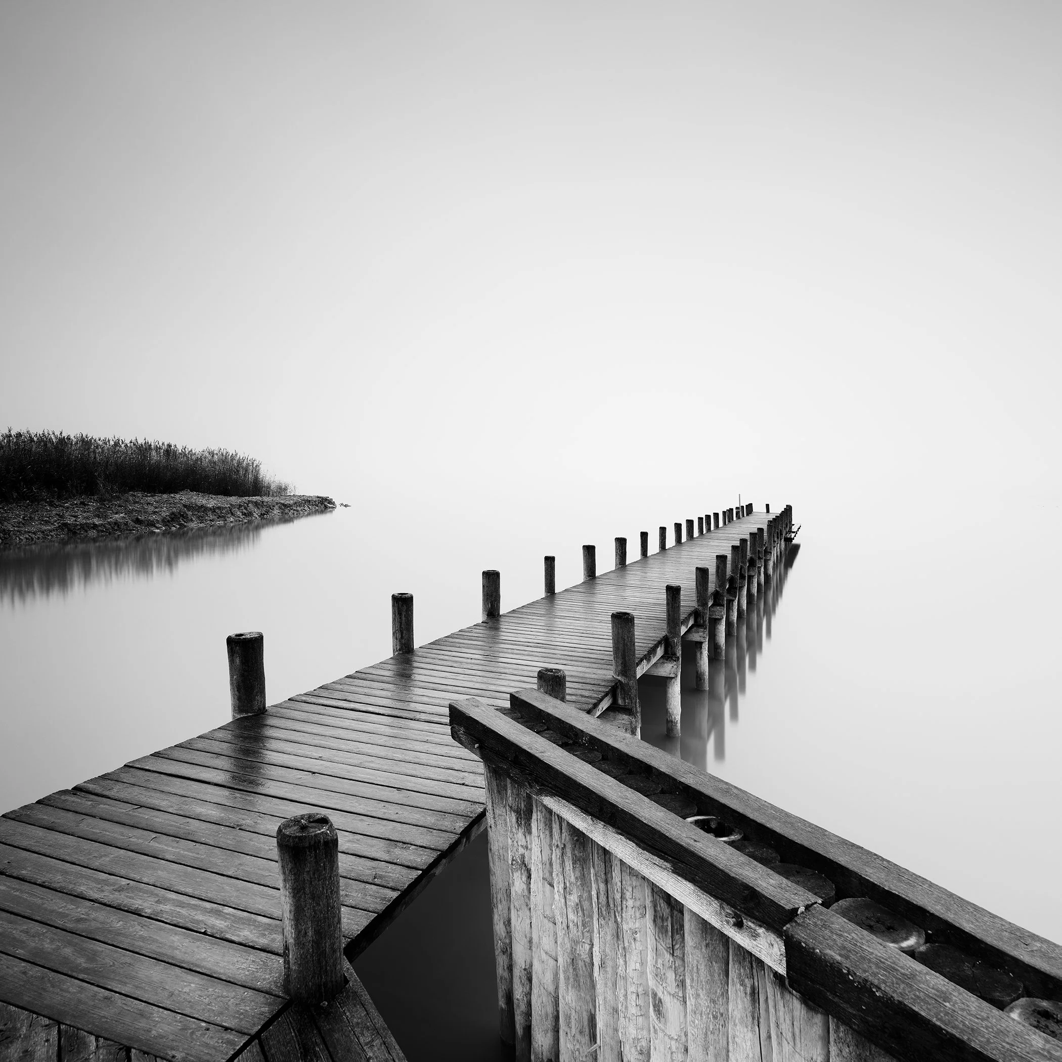 Gerald Berghammer - Black and white photography. Wooden pier extending out over calm water, with a foggy background and some reeds on the shoreline.