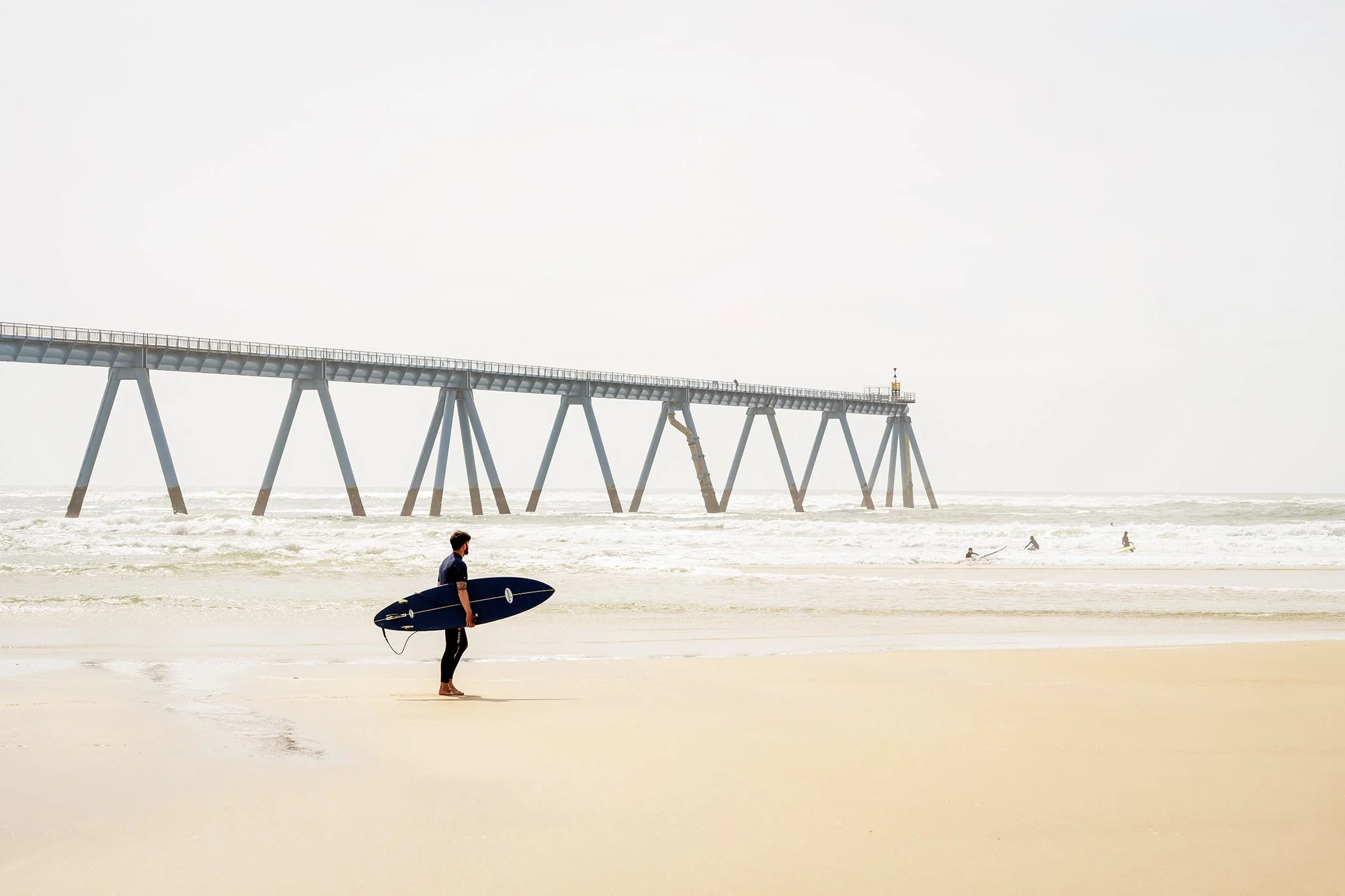 Wave Watcher Study 2 by Gerald Berghammer, minimalist colour fine art photograph of a solitary surfer, sandy beach, ocean waves and pier in France.