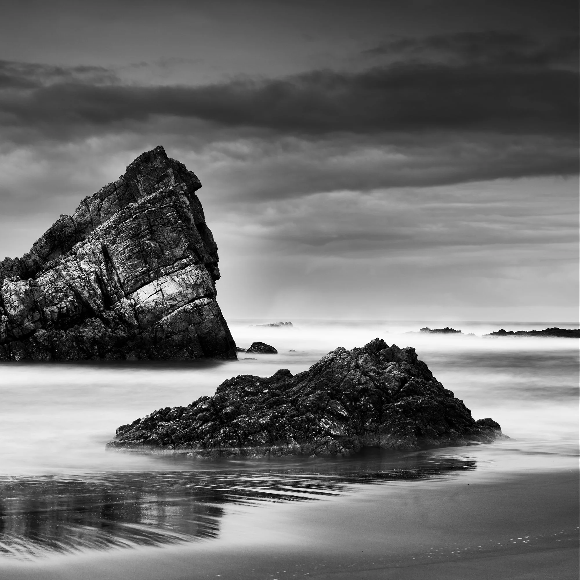 © 2023 Gerald Berghammer. Black-and-white photograph of jagged sea rocks emerging from calm water under a cloudy sky. Print detail 2