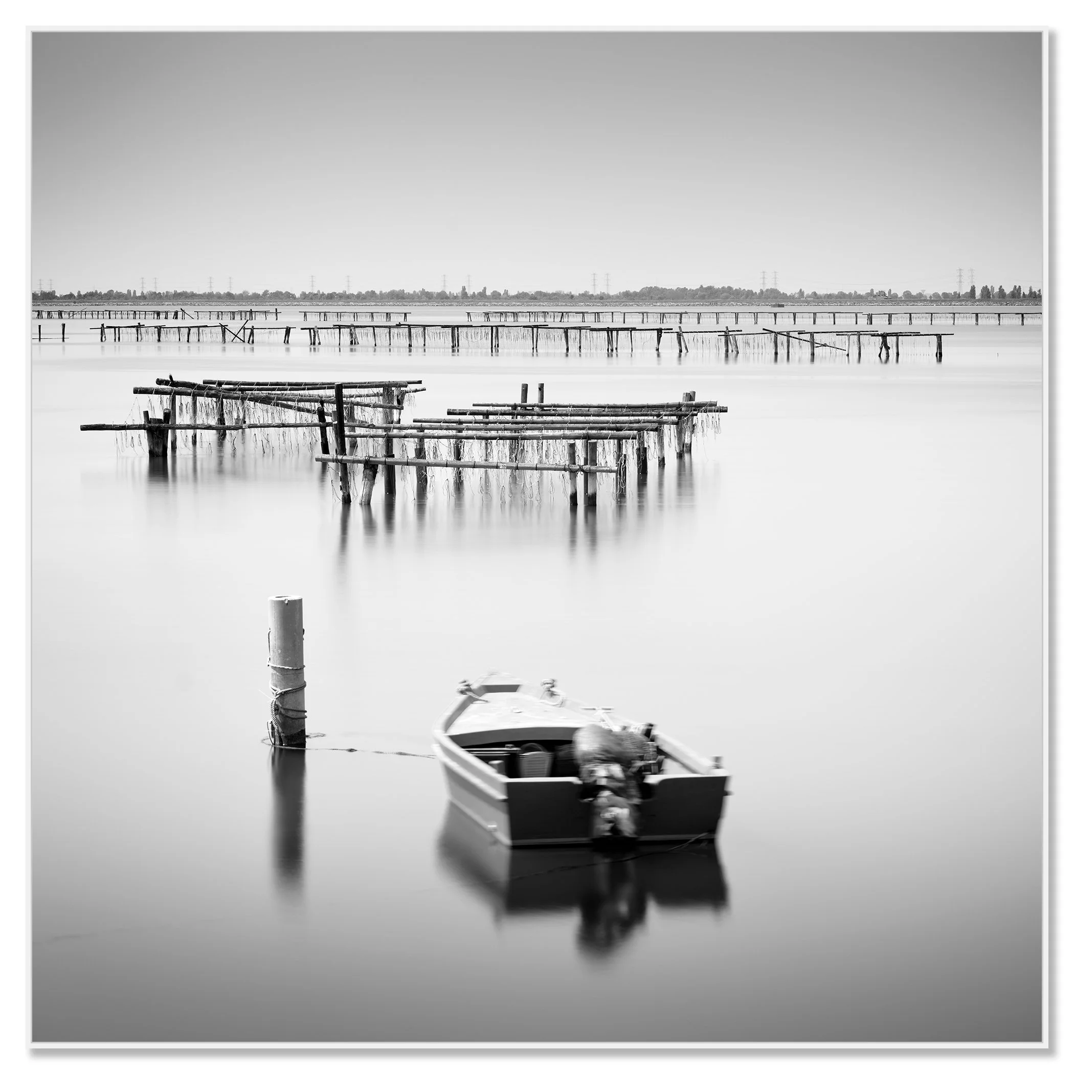 Black-and-white seascape photograph of a small boat tied to a post on calm water, with wooden aquaculture piers in Italy – framed ArtBox white