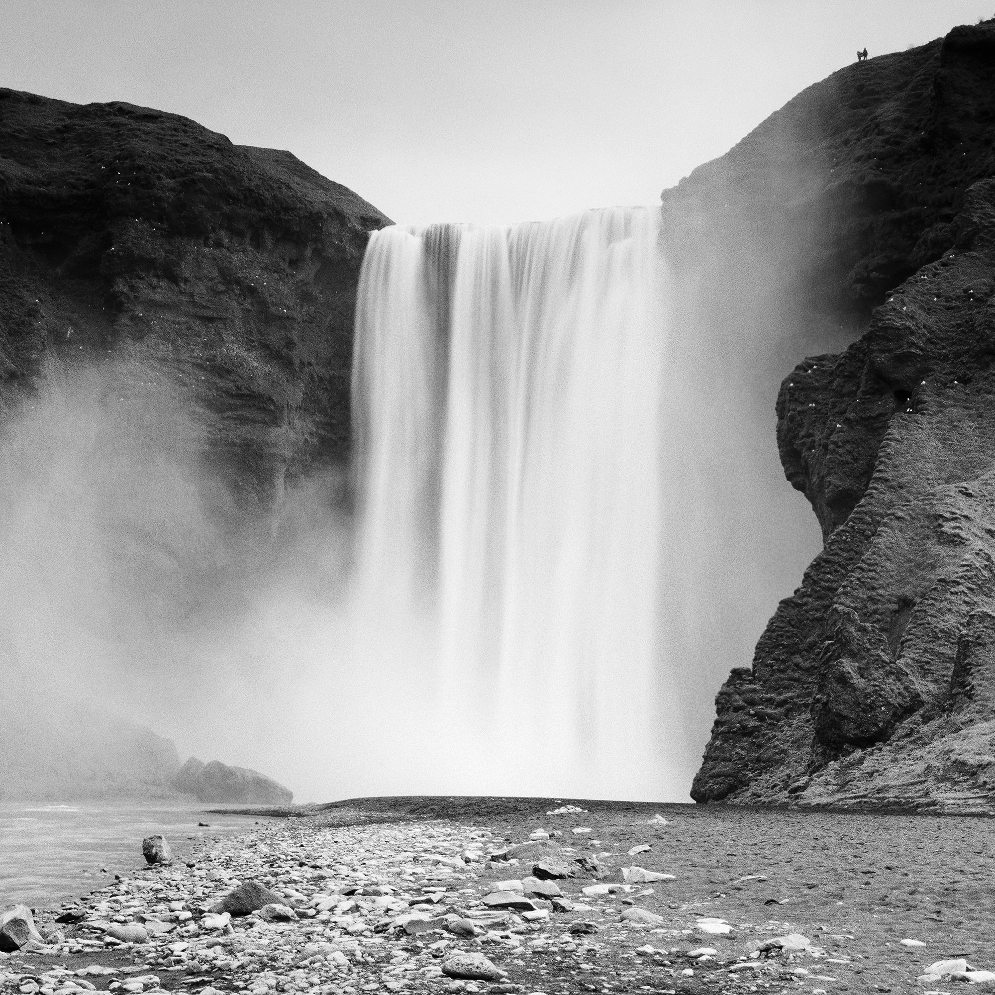 Gerald Berghammer - Black and white long exposure waterscape photography. A large waterfall flowing between rugged cliffs into a rocky river. Print detail 3