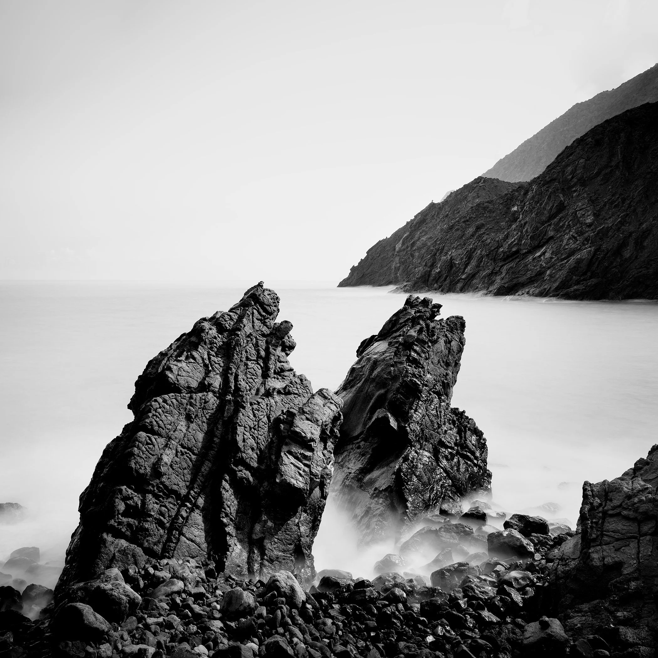 Black and white seascape photograph of a rocky Atlantic shoreline with large boulders in the water and mountains in the background.