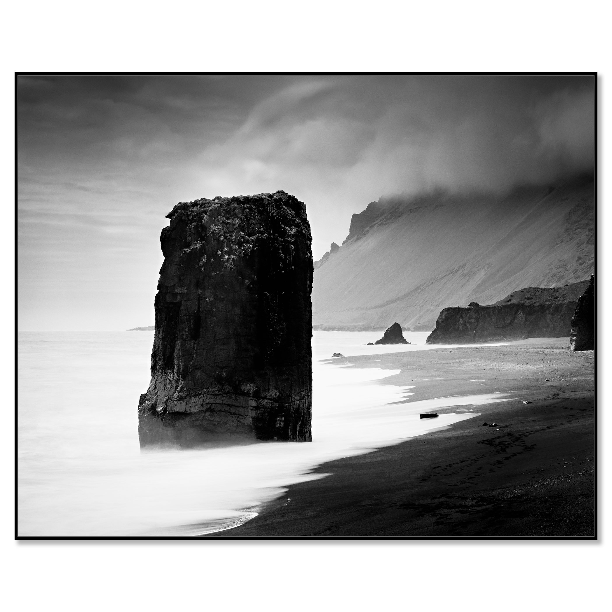 Black-and-white photograph of a rock monolith in the sea, with smooth long-exposure water, dark sand, and misty cliffs, framed ArtBox black