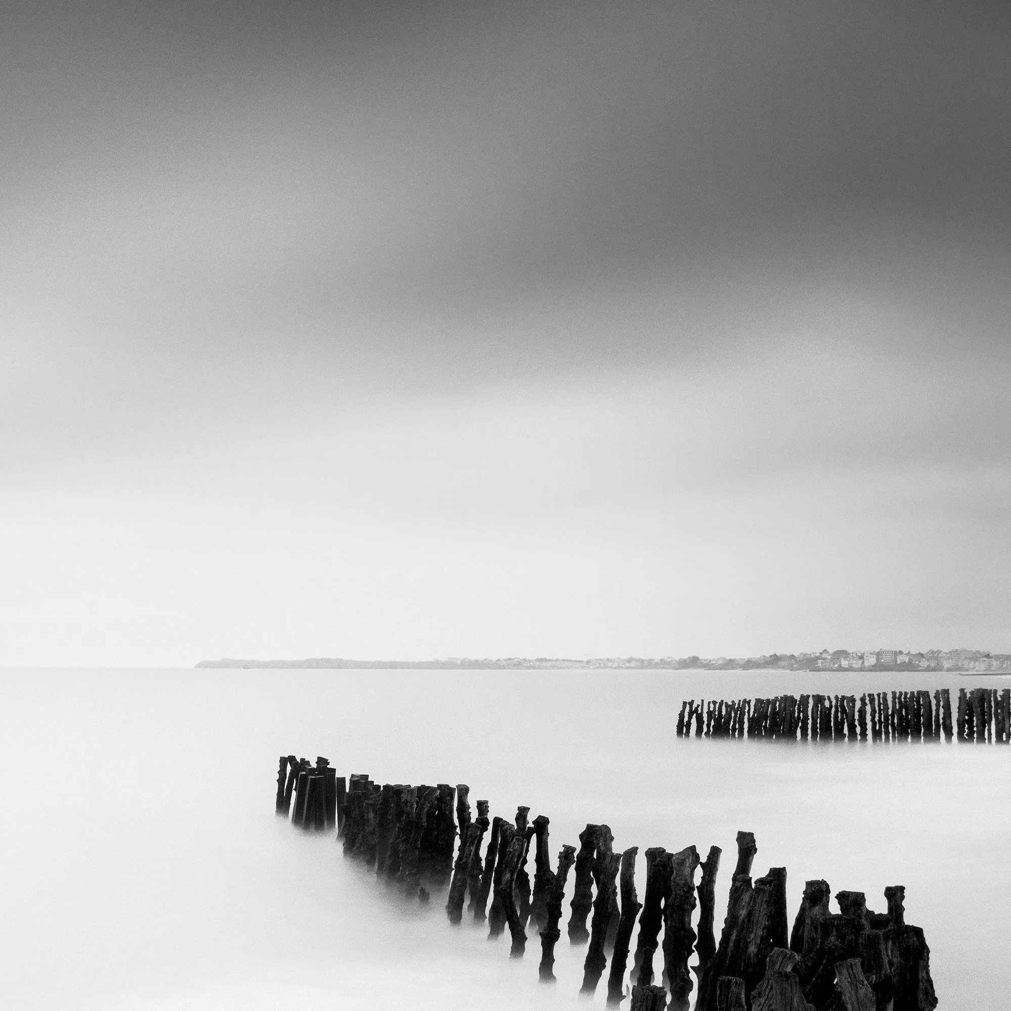 Cropped view of Sea Breakers with wooden groynes, calm water and the distant French Atlantic coastline by Gerald Berghammer.