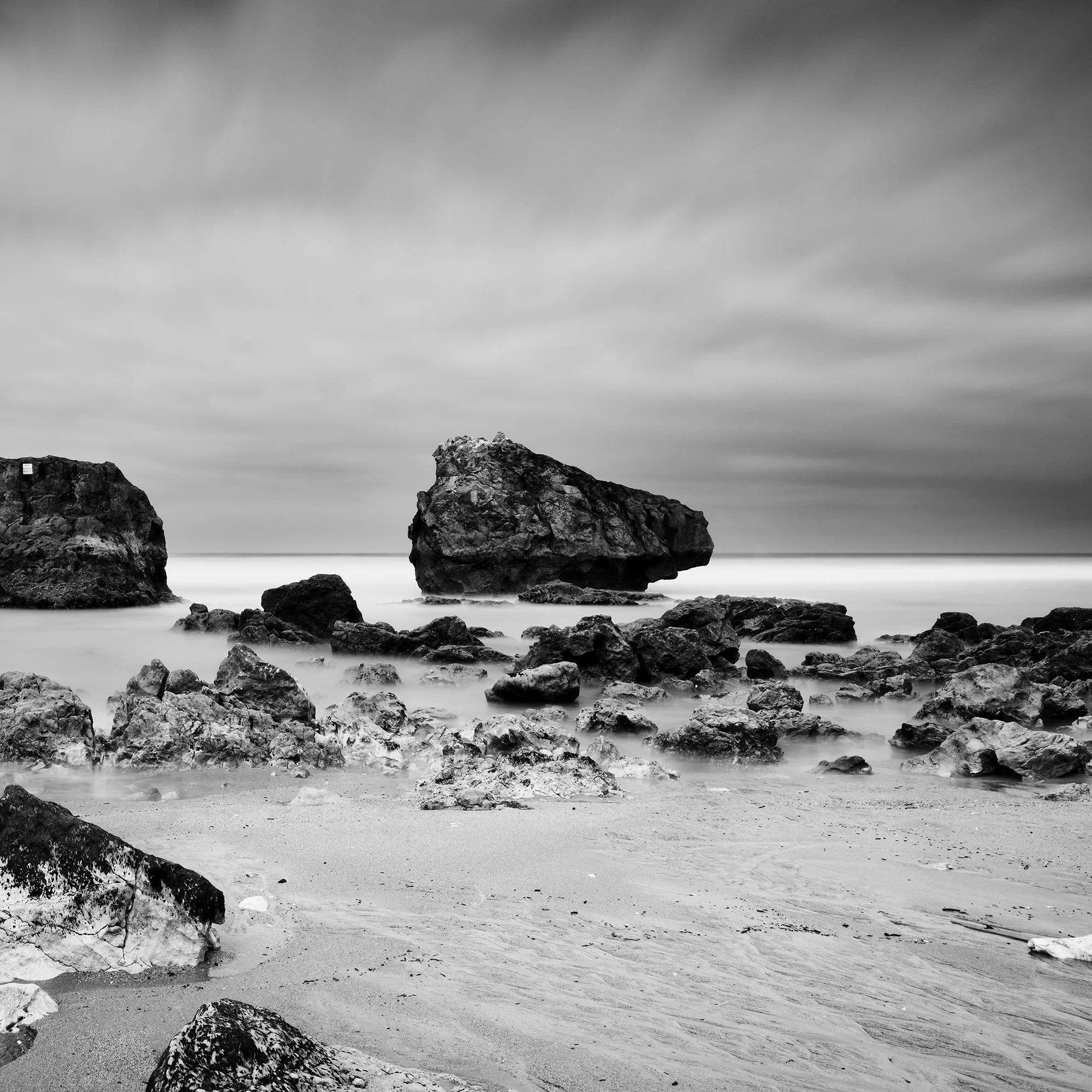 Gerald Berghammer - Black and white landscape photo. Rocky beach with two large rocks protruding from the water, cloudy sky, and smooth ocean surface. Print detail 2