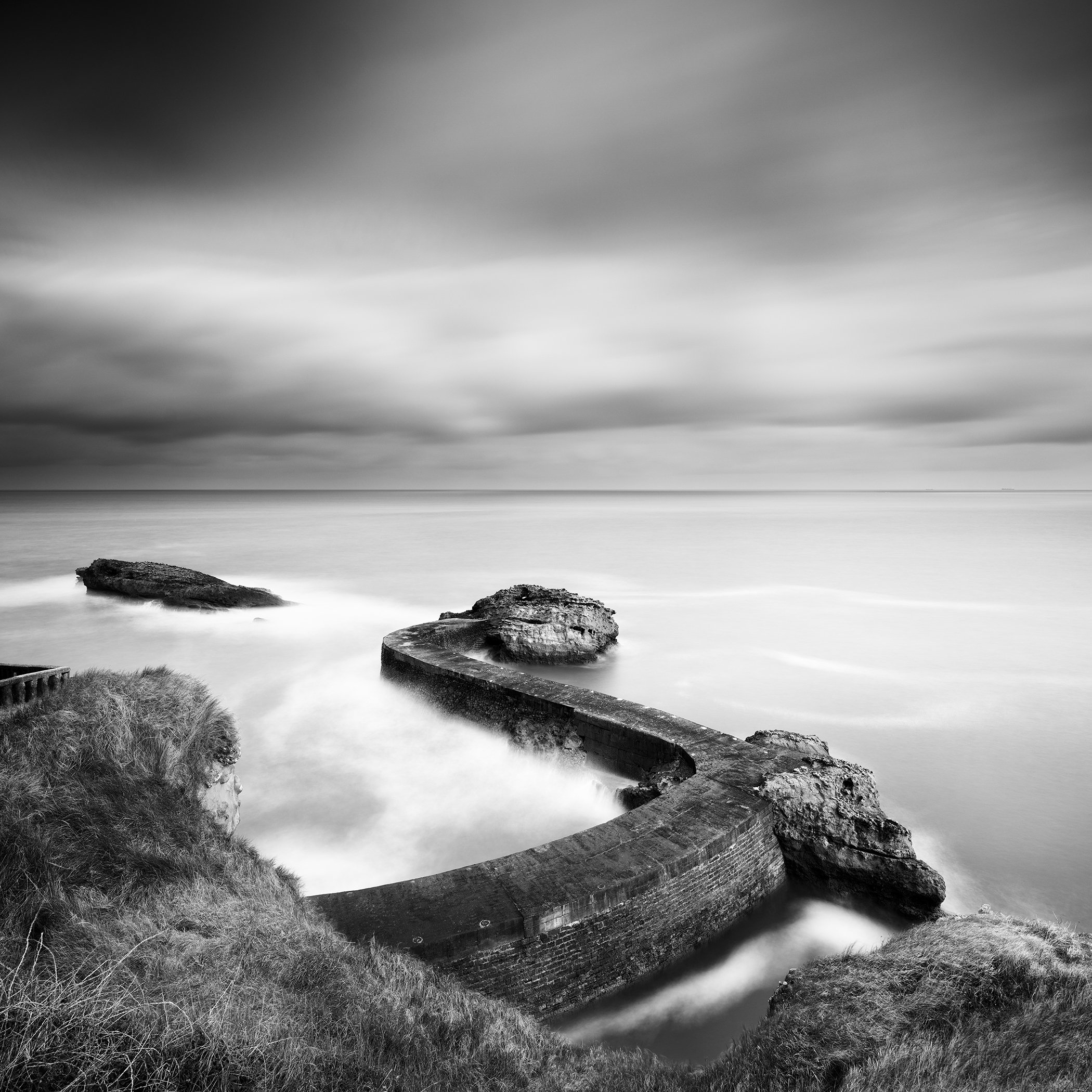 Monochrome coastal scene showing a curved stone breakwater pier extending into a calm sea beneath dramatic clouds