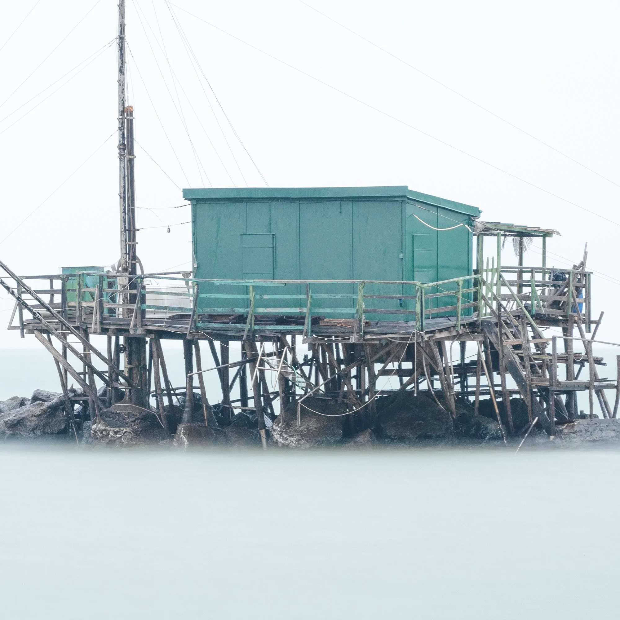 © 2024 Gerald Berghammer - Color minimalist photography. A small house built on stilts over water on rocks, with a fishing pole, in a minimalist, misty seascape. Print detail 3