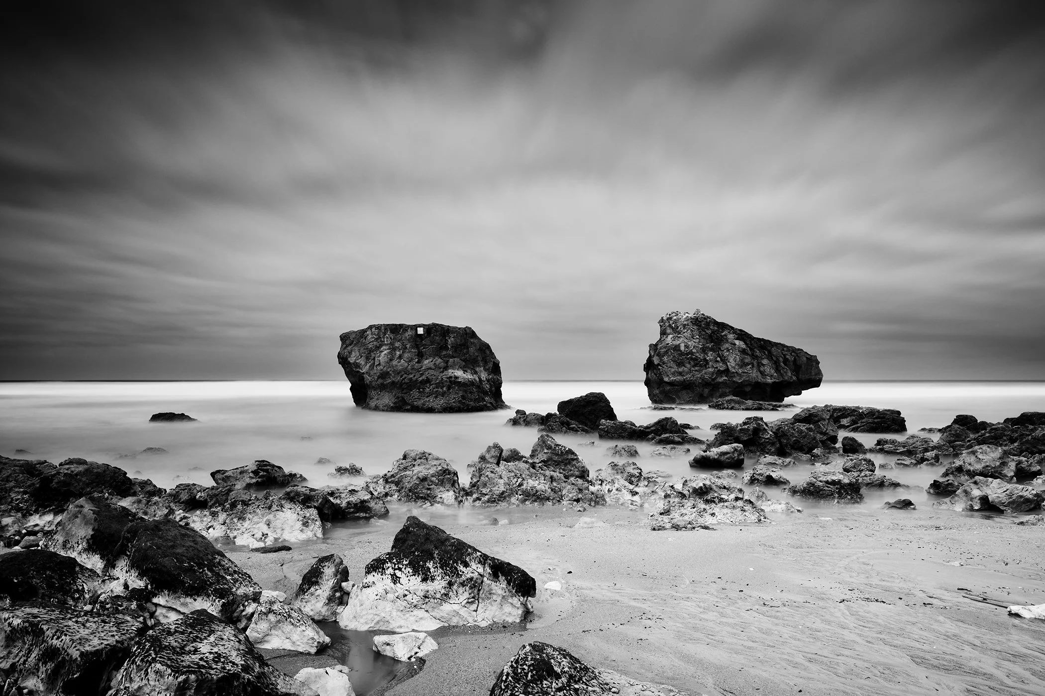 Black and white coastal landscape with two large rocks in the sea, cloudy sky and calm, smooth water