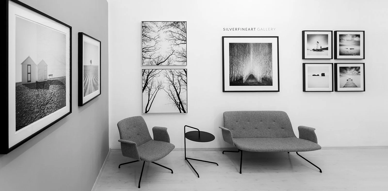 A grayscale photo of the SILVERFINEART gallery with framed black and white photographs on the walls, including landscapes and trees, and modern chairs and a small table in the foreground.