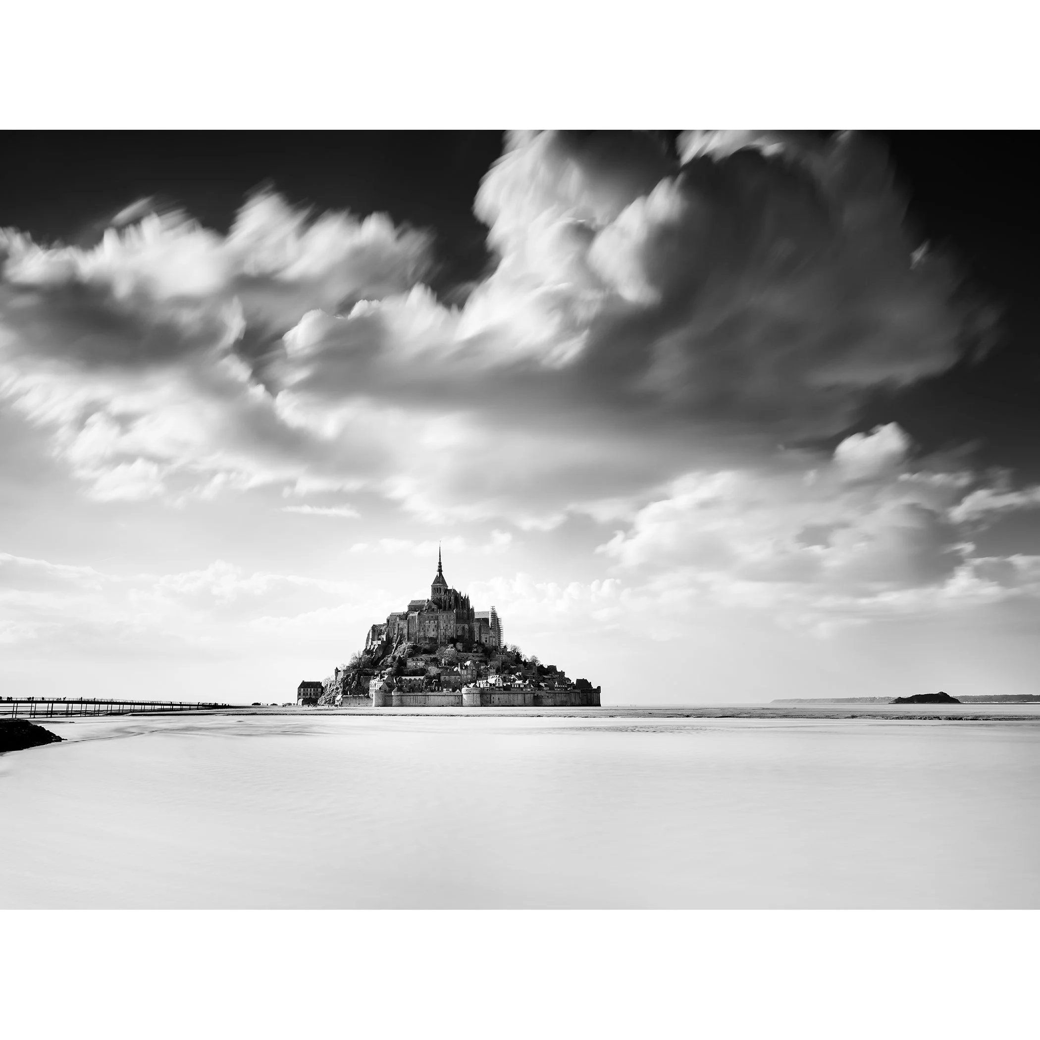 © 2023 Gerald Berghammer - B&W photography. Mont Saint-Michel, a historic island with a tall spire and medieval architecture, surrounded by water and clouds.