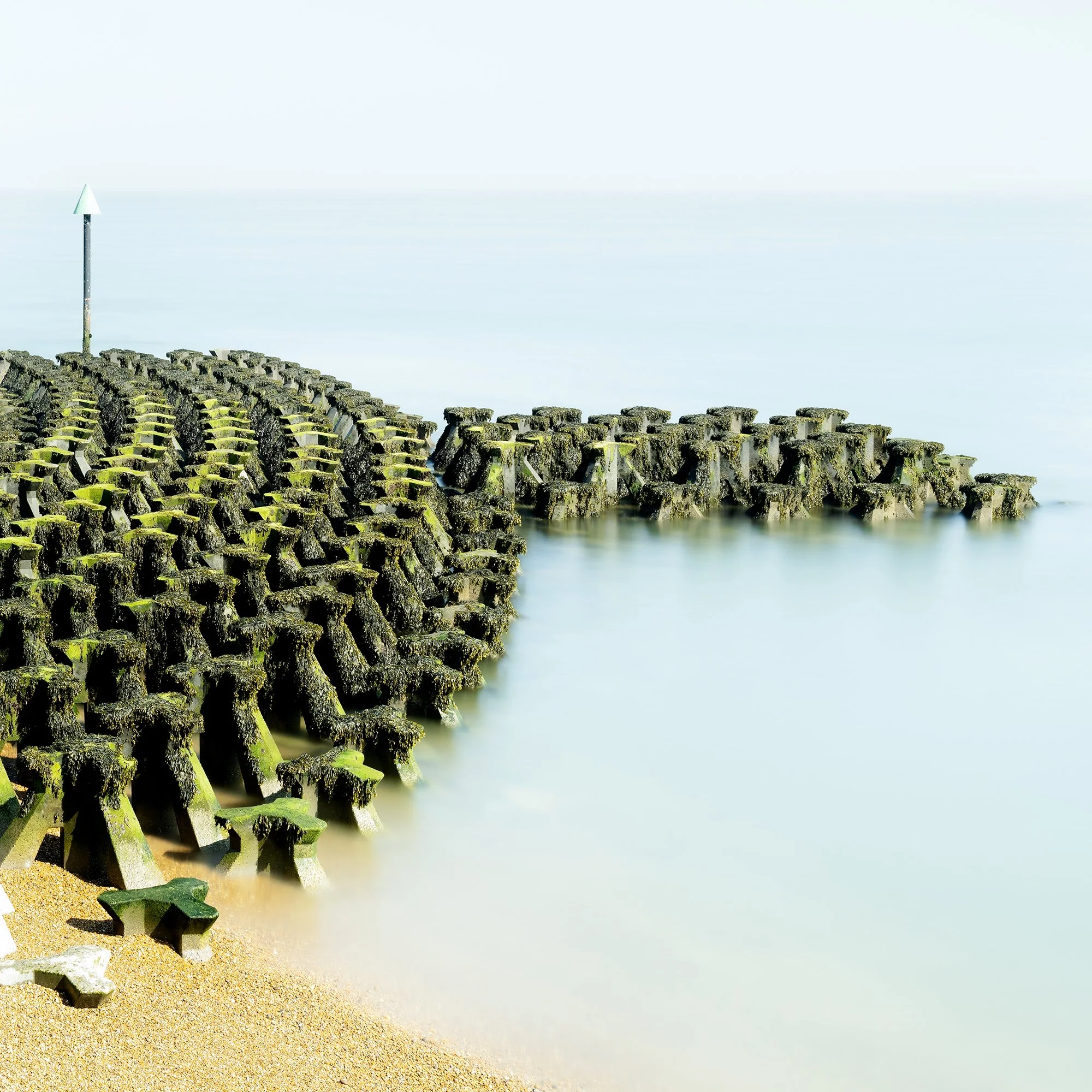 Gerald Berghammer - Color seascape panorama photography. Concrete breakwater structures on a beach extending into the water, with a pole at the end. Print detail 2