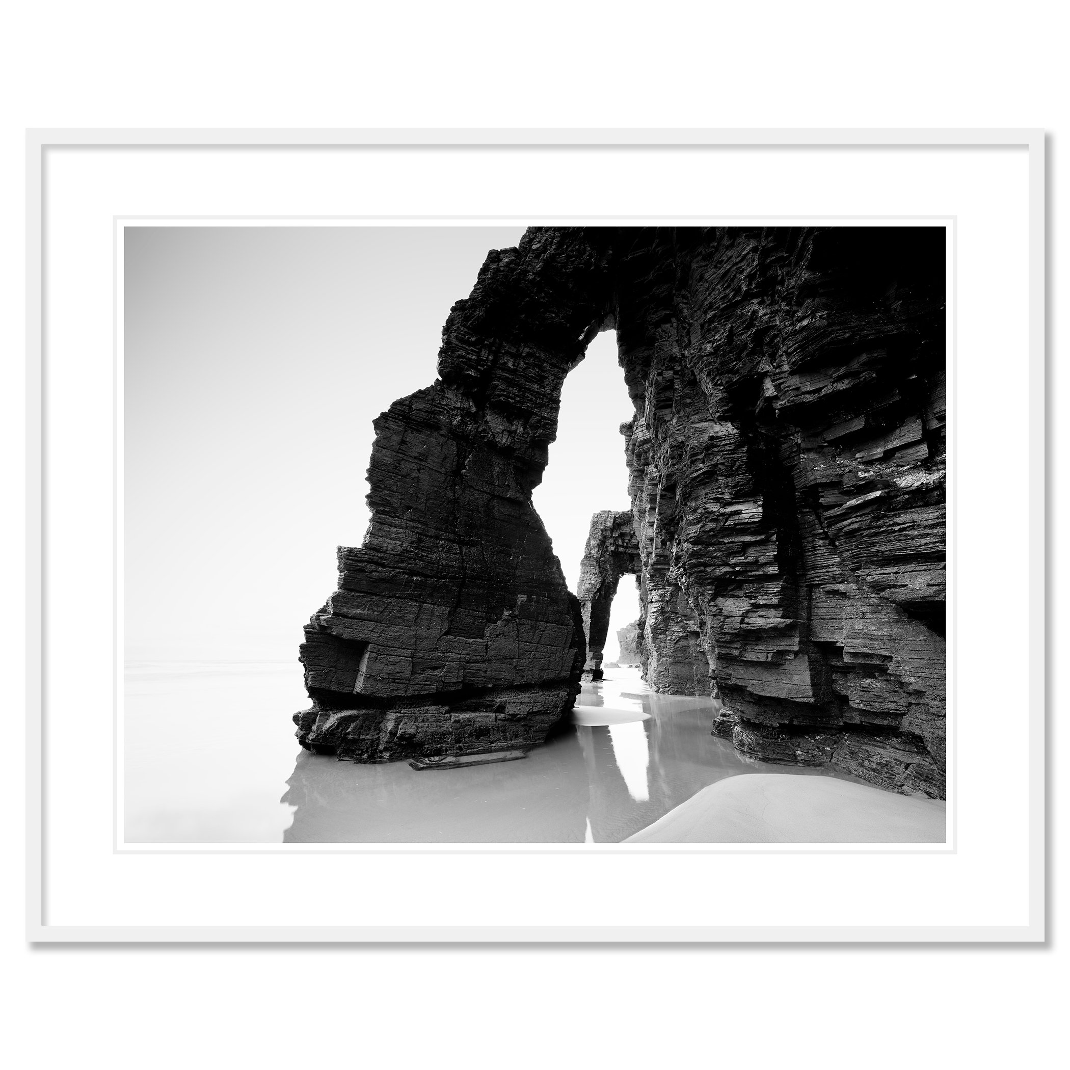 Gerald Berghammer - Black and white beach seascape photography. Large natural rock arch formations on a beach with water reflecting the rocks. Classic framed white