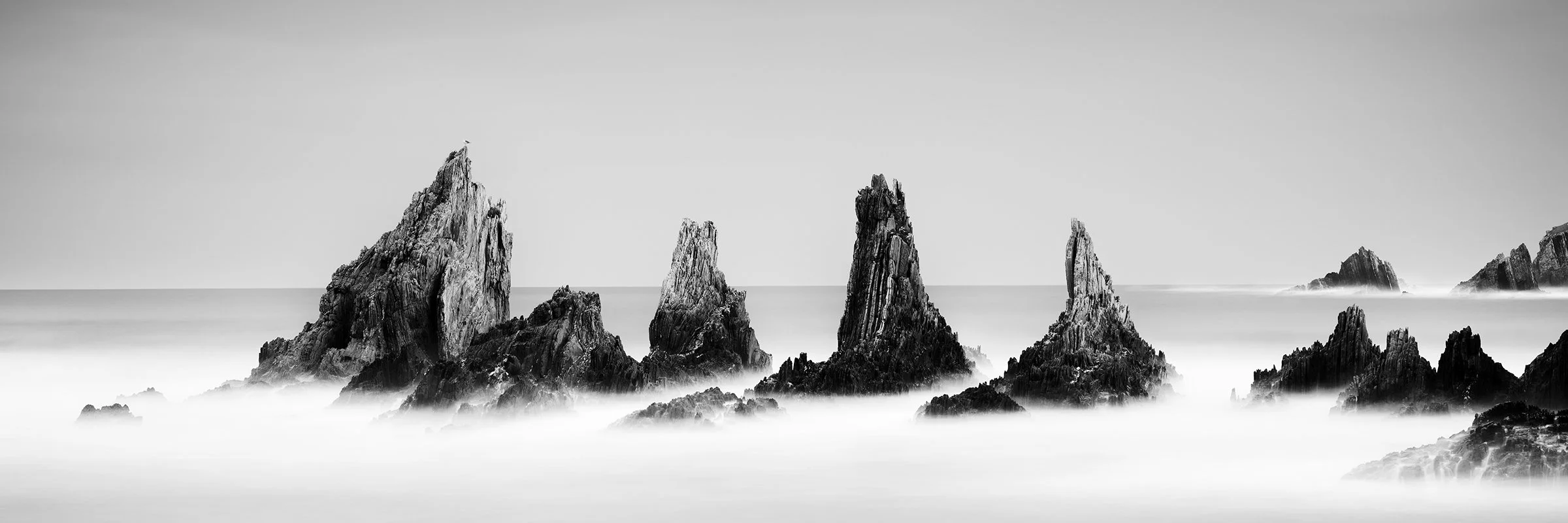 Rocky Peaks Panorama Study 3 by Gerald Berghammer, black-and-white long-exposure photograph of jagged sea stacks rising from misty Atlantic water in Spain.