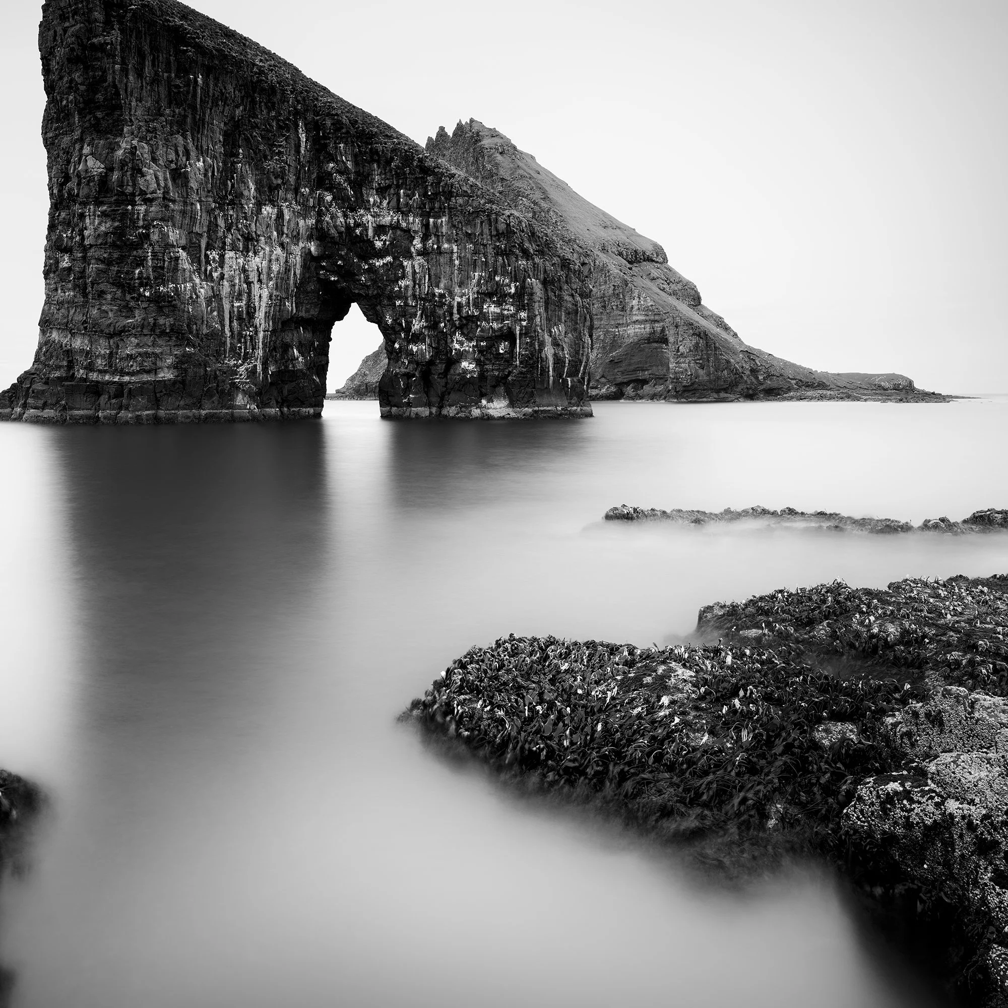 Black and white coastal landscape with a striking sea stack arch and soft long-exposure water – cropped view